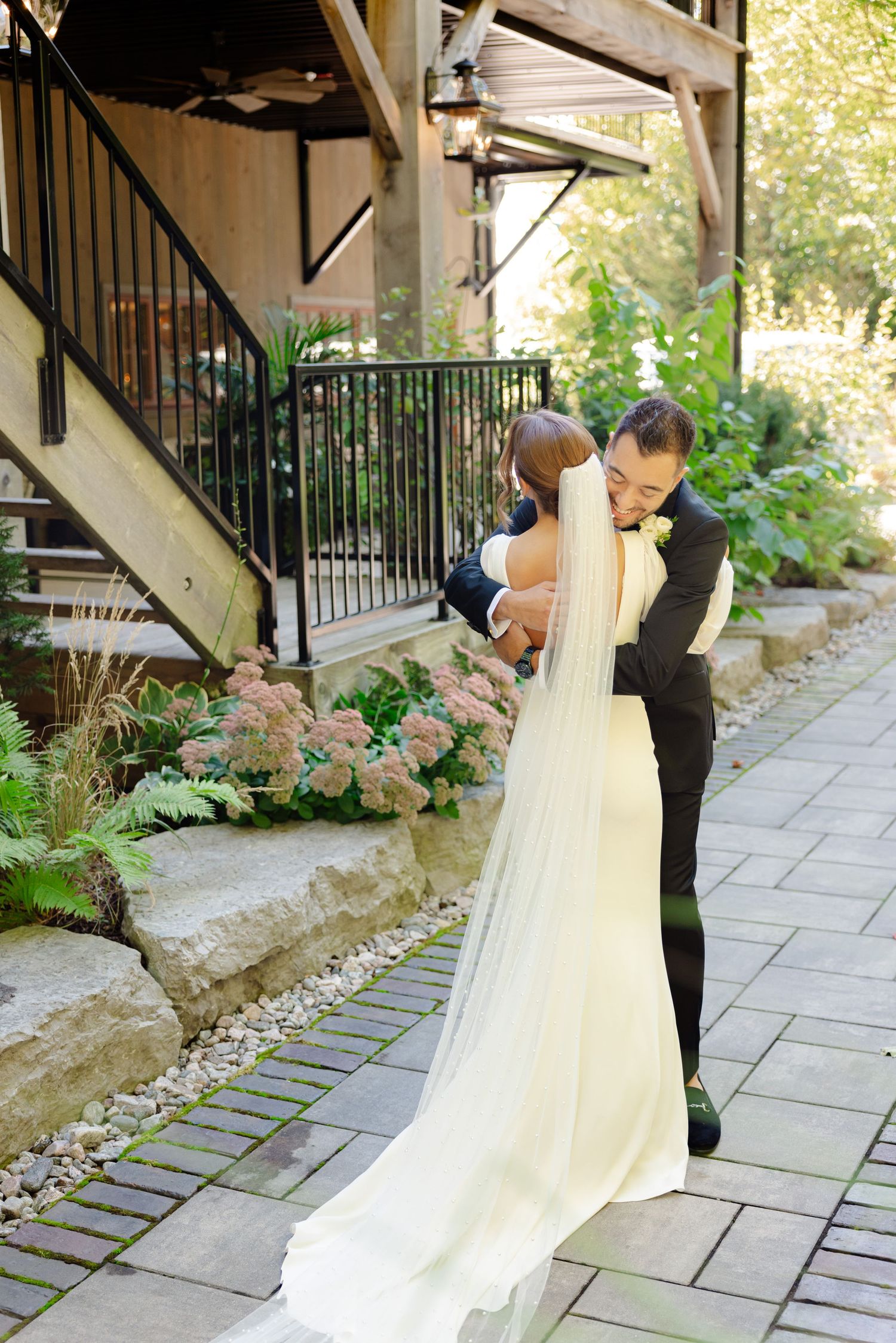 Wedding couple embraces on a garden path beside decorative stone walls and flowering plants.