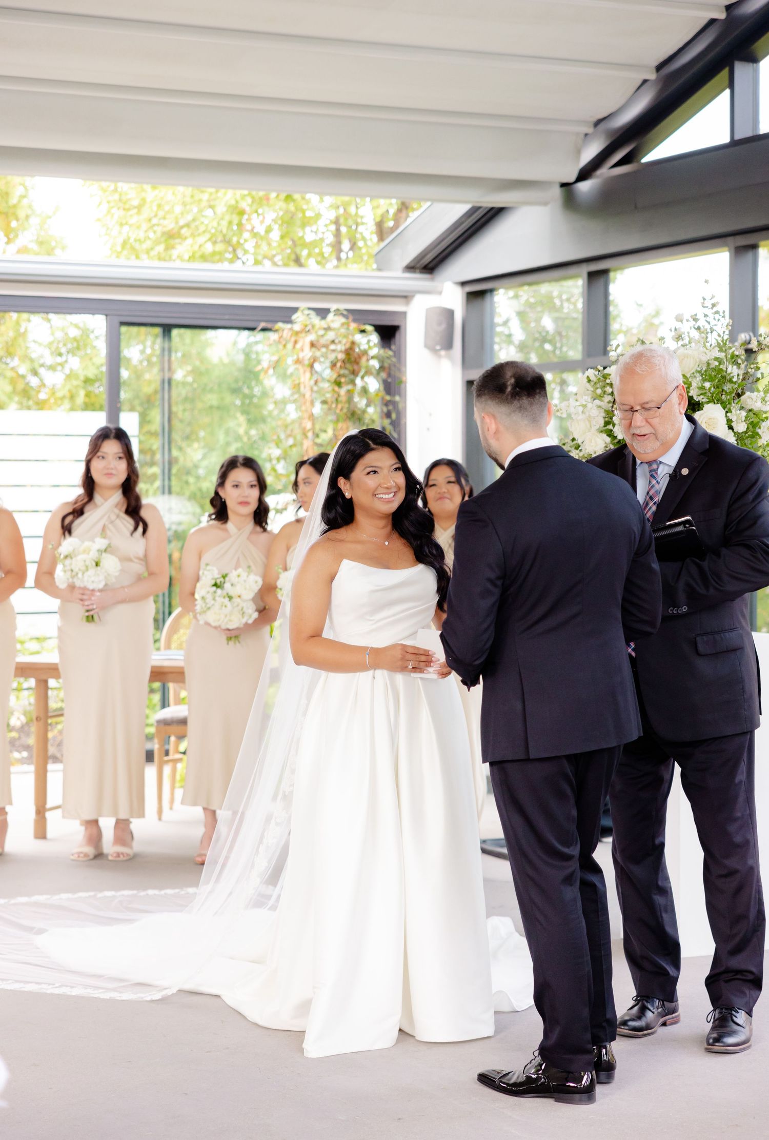 A wedding ceremony in progress with bridesmaids in blush dresses and white floral arrangements in a bright glass venue.