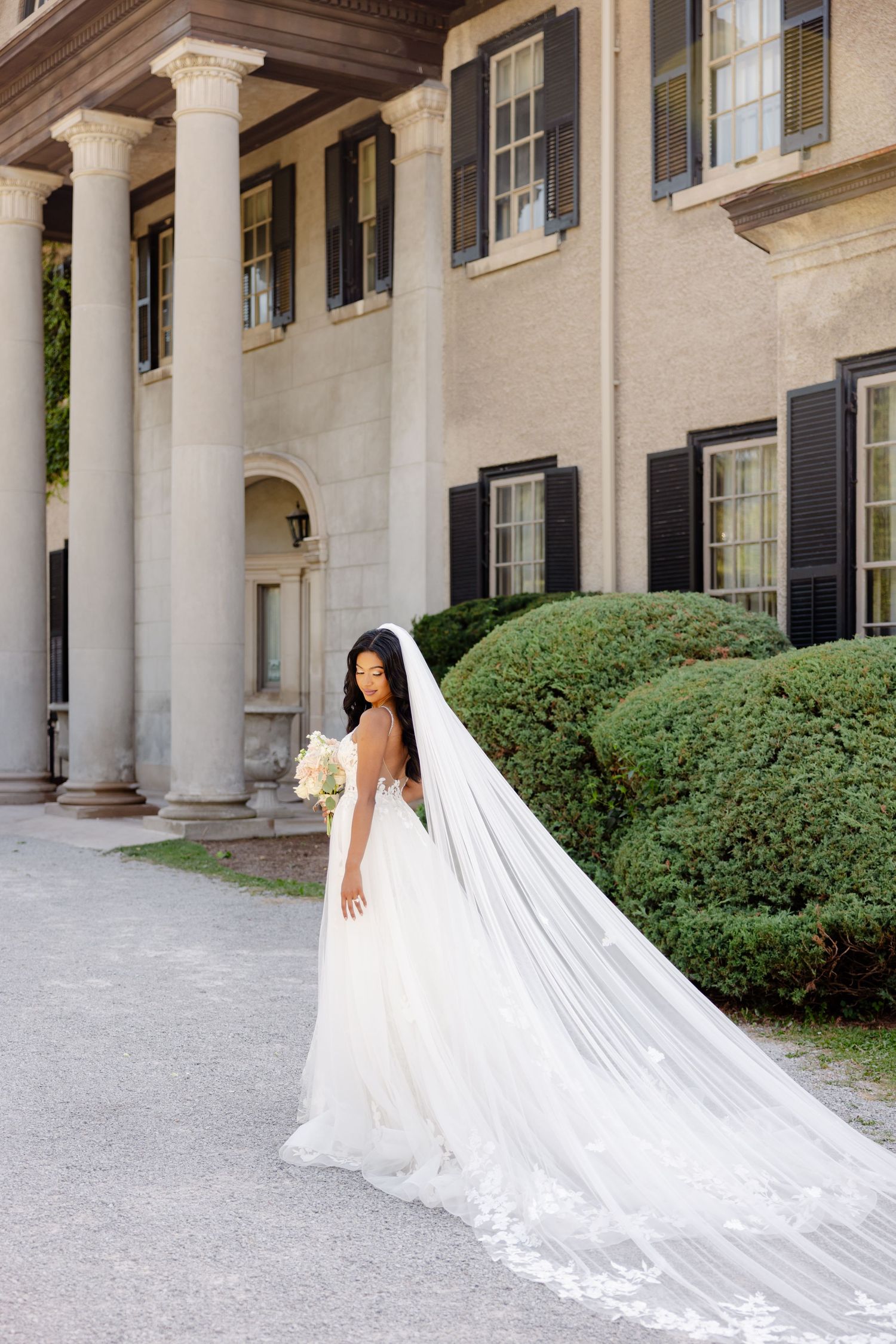 A stunning bride poses in a white lace gown with cathedral veil against a stately mansion with white columns.