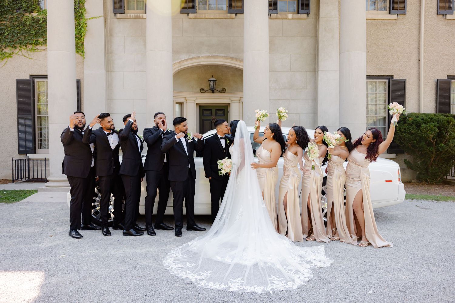Wedding party in formal attire poses outside elegant building with columns and white cathedral veil trailing behind.