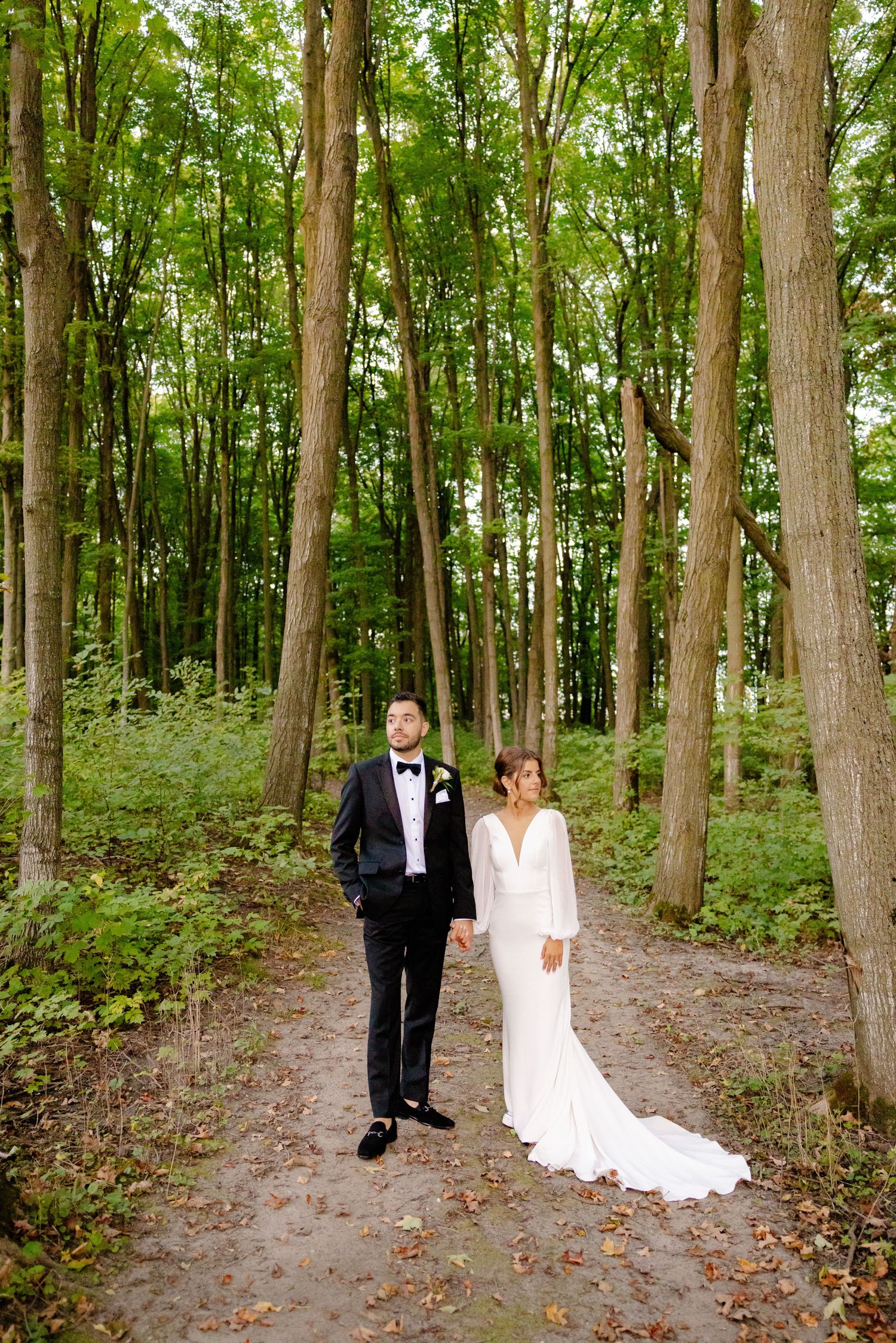 A wedding couple stands on a forest path surrounded by tall trees.