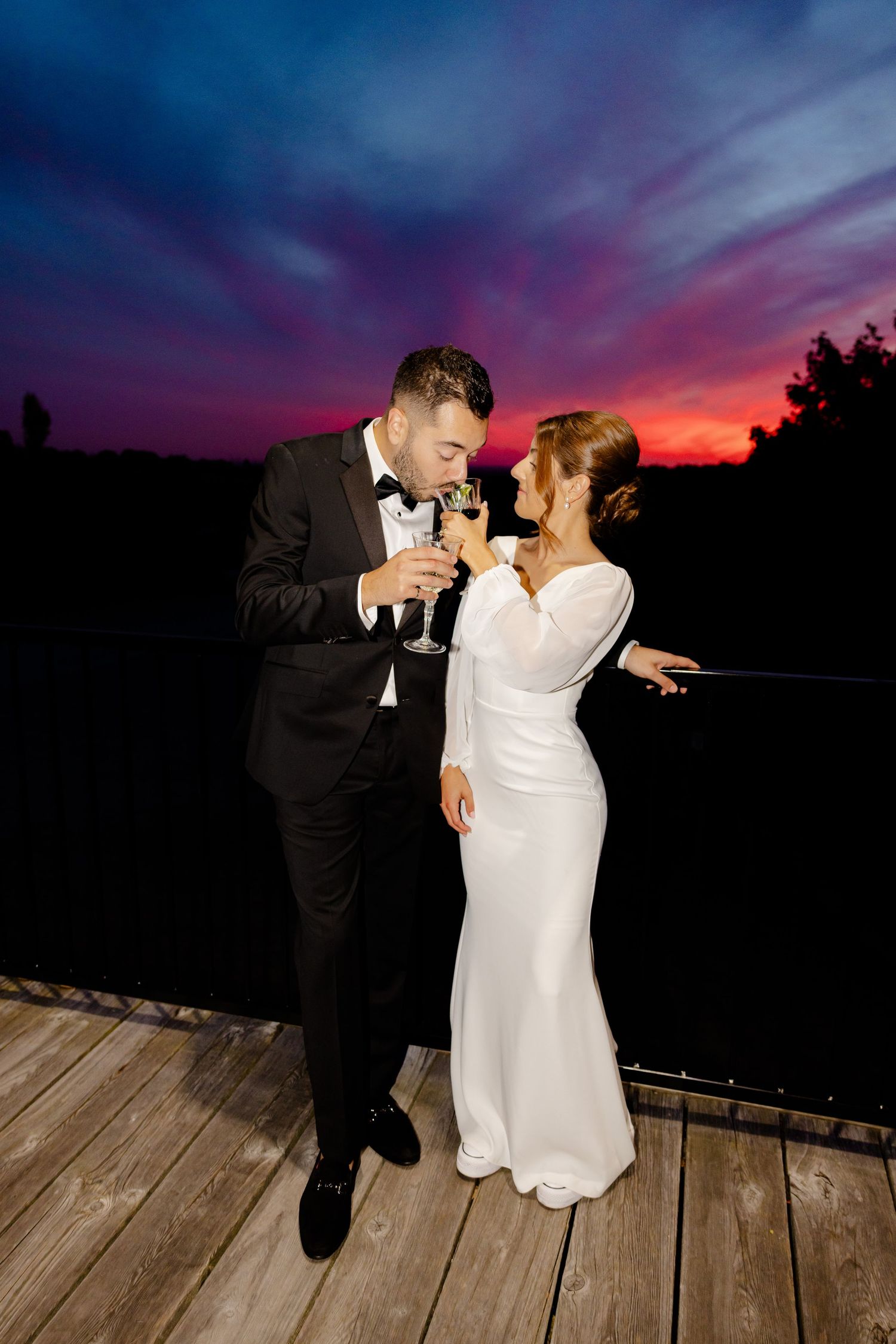 A bride and groom sharing a playful moment with champagne on a wooden deck at dusk.