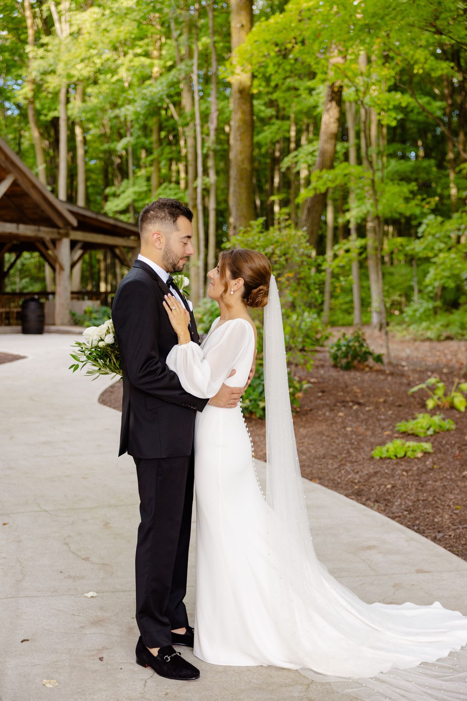 Elegant bride and groom share an intimate moment on a winding forest path near a wooden pavilion.