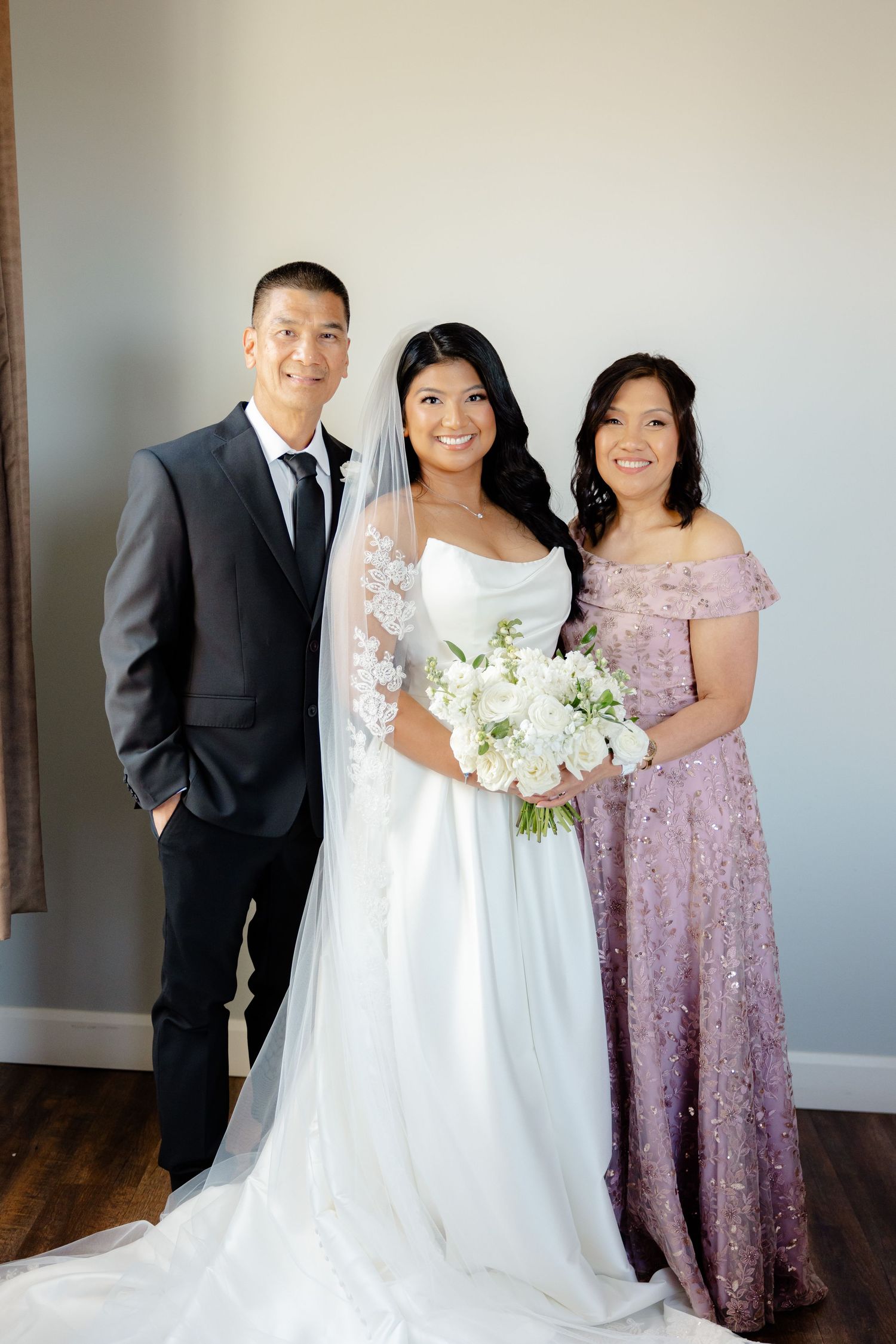 Wedding party portrait with bride in white gown flanked by formal-dressed companions against neutral wall.