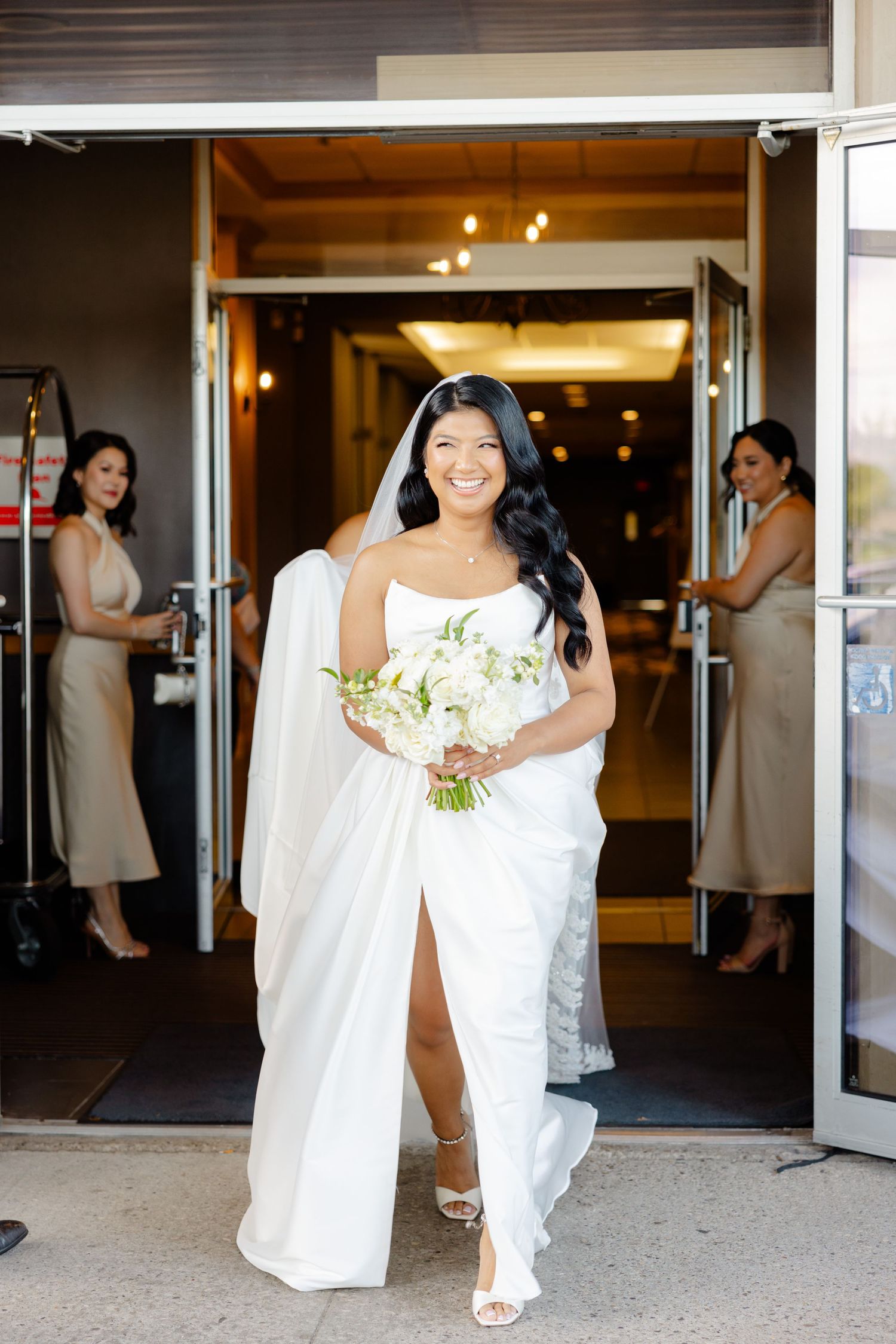 Bride in a white wedding gown enters through glass doors carrying a white floral bouquet.