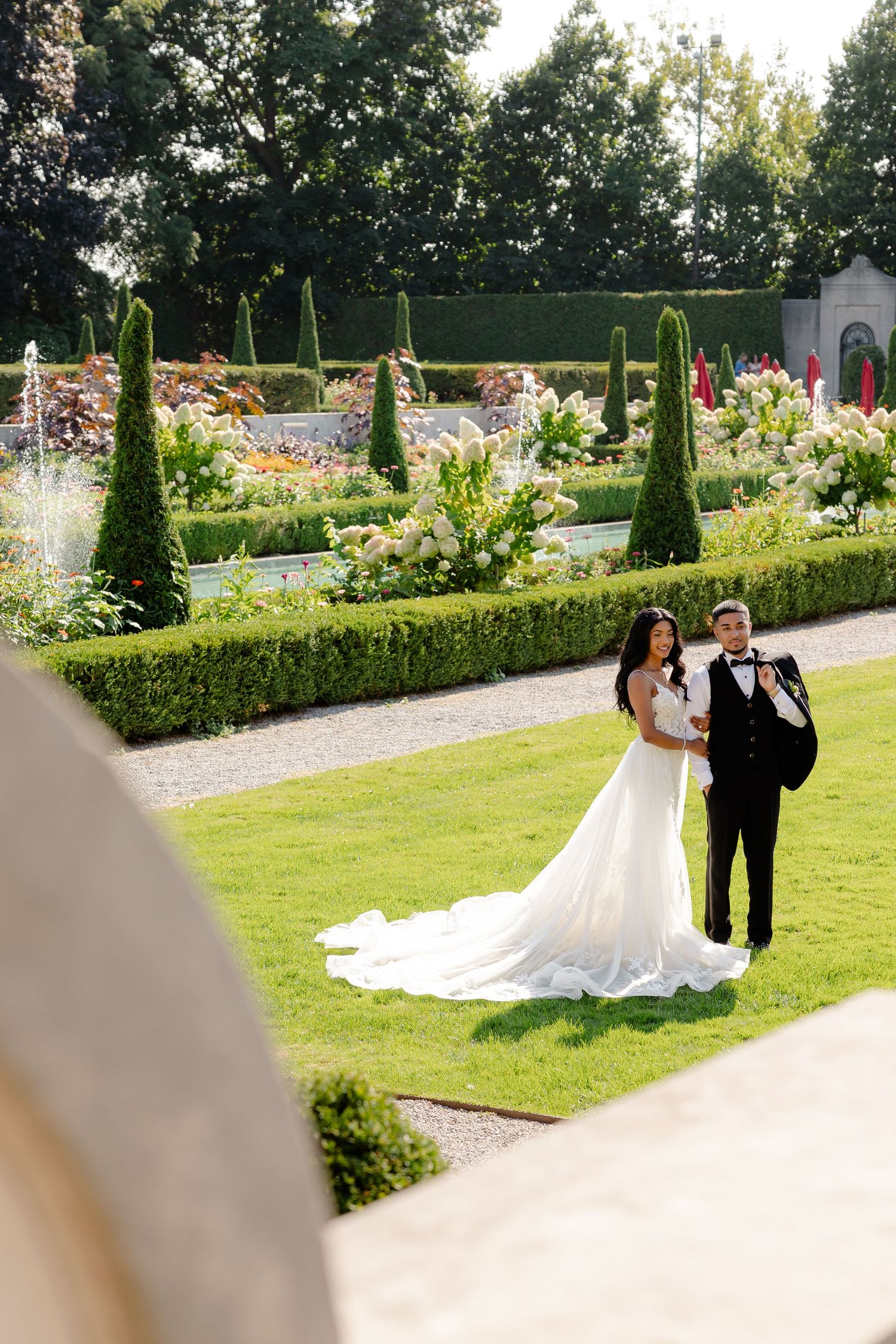 A couple poses on manicured lawn surrounded by formal garden with topiary and flowers.