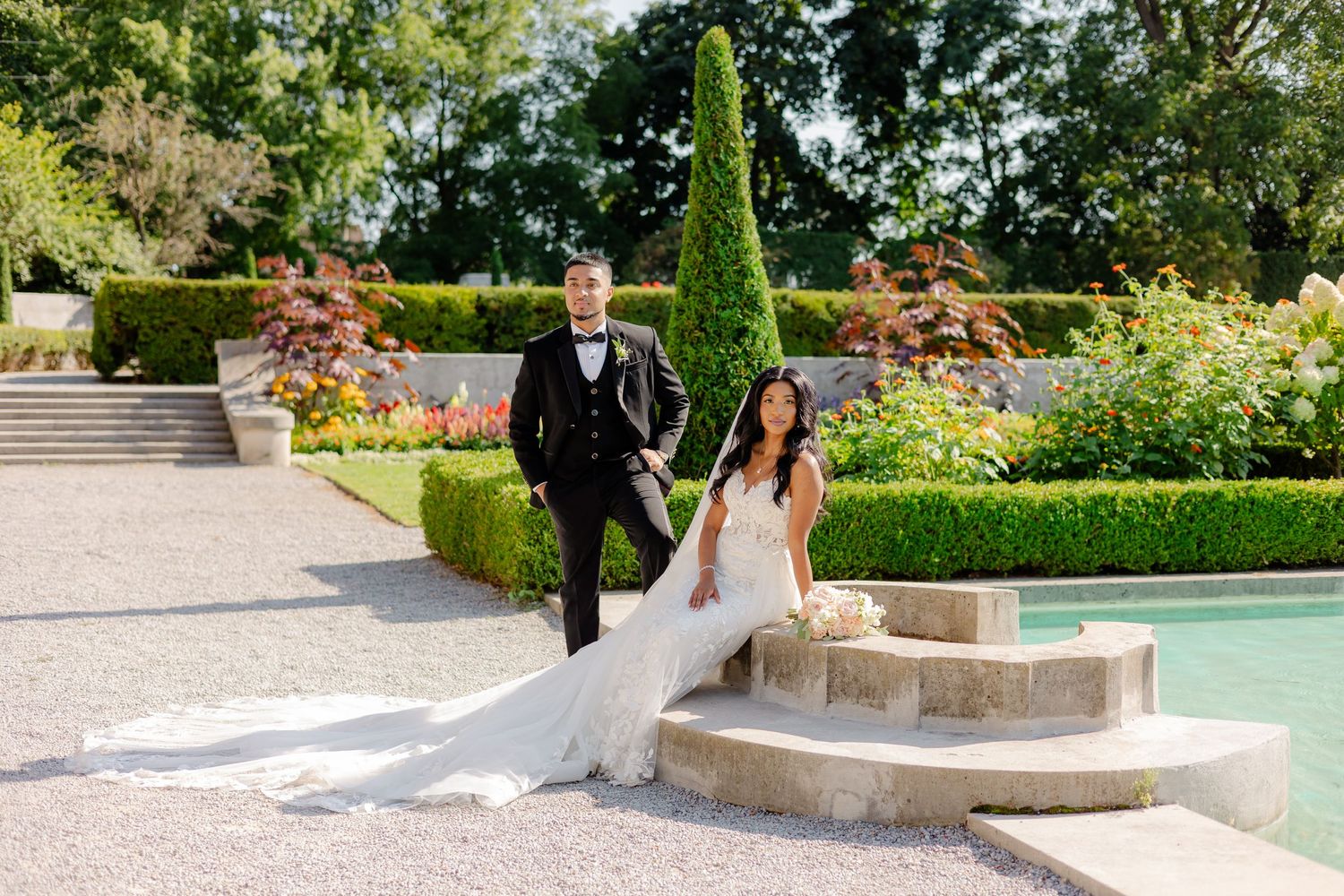 Couple in wedding attire walk together through a landscaped garden with stone steps.