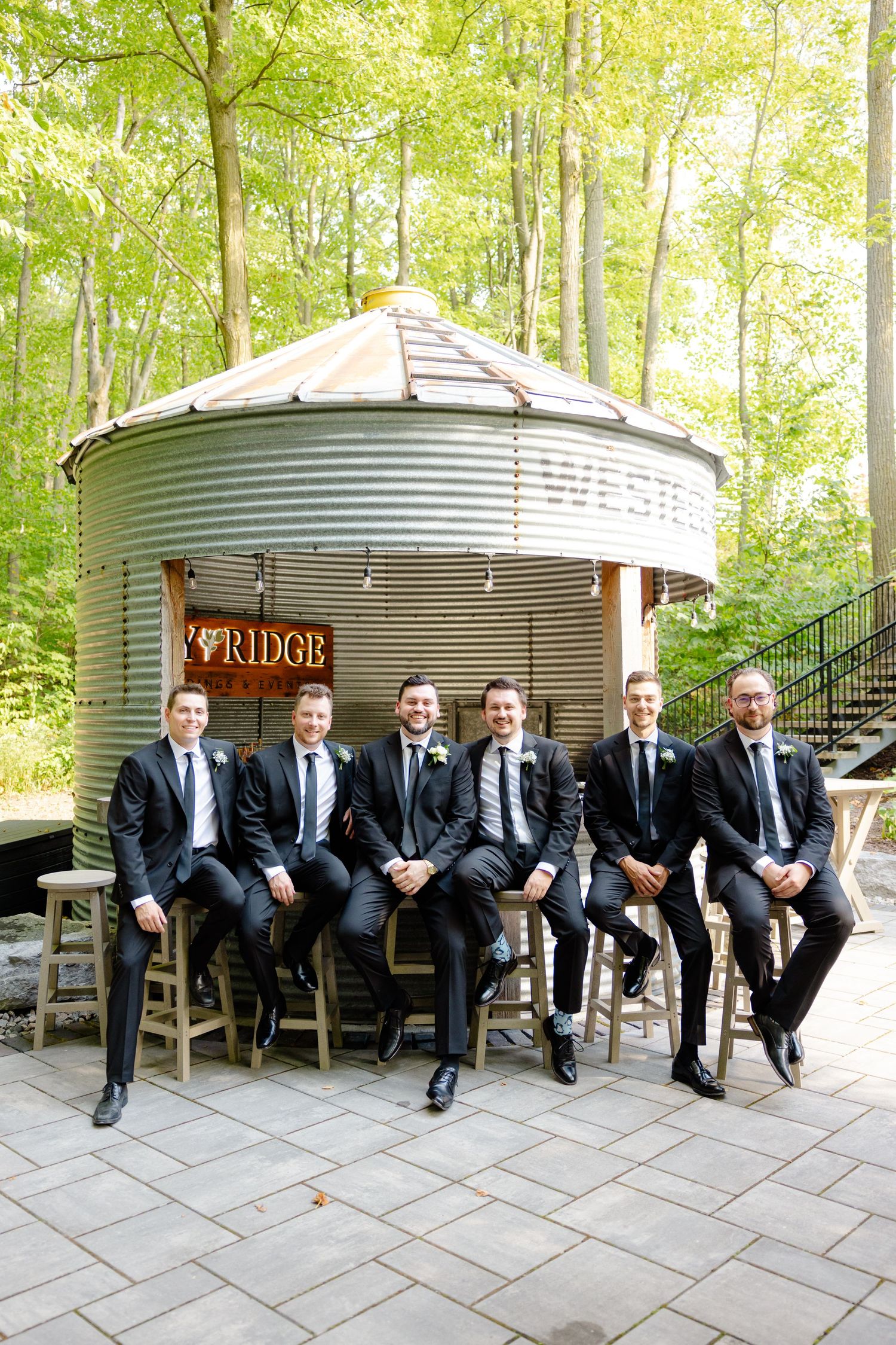 Groomsmen group seated on bar stools in front of metal silo structure with Ivy Ridge sign.