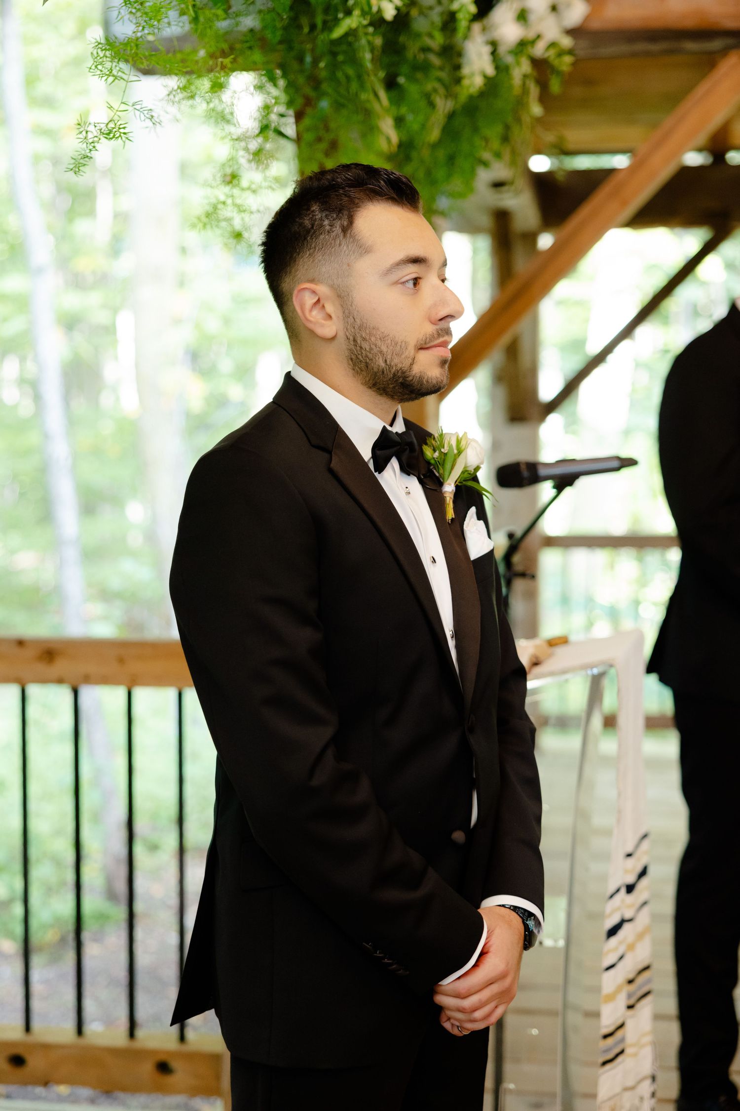 Groom in black tuxedo standing at altar in outdoor wooden pavilion.