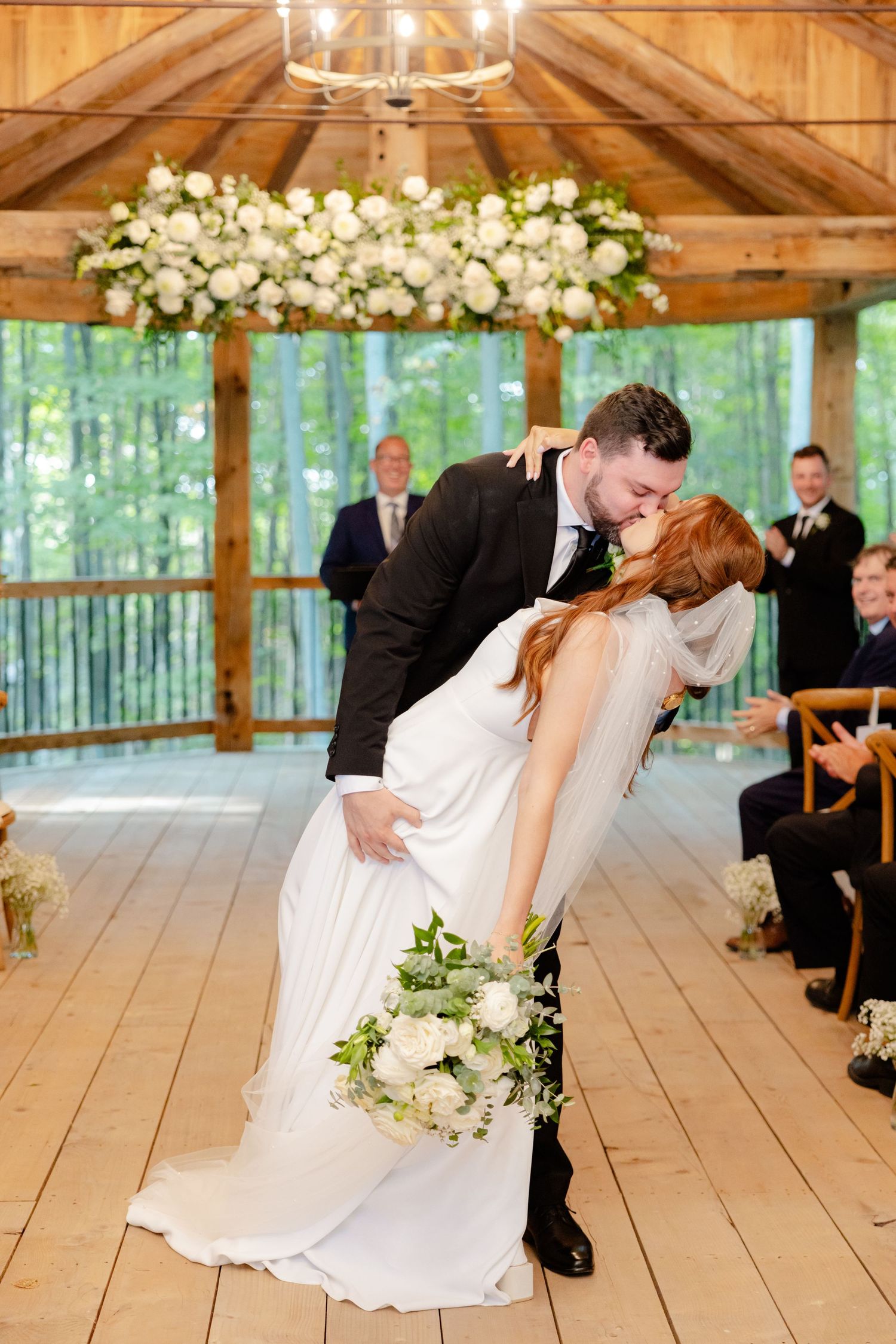 A romantic dip kiss during a wedding ceremony under a floral chandelier in a wooden pavilion.
