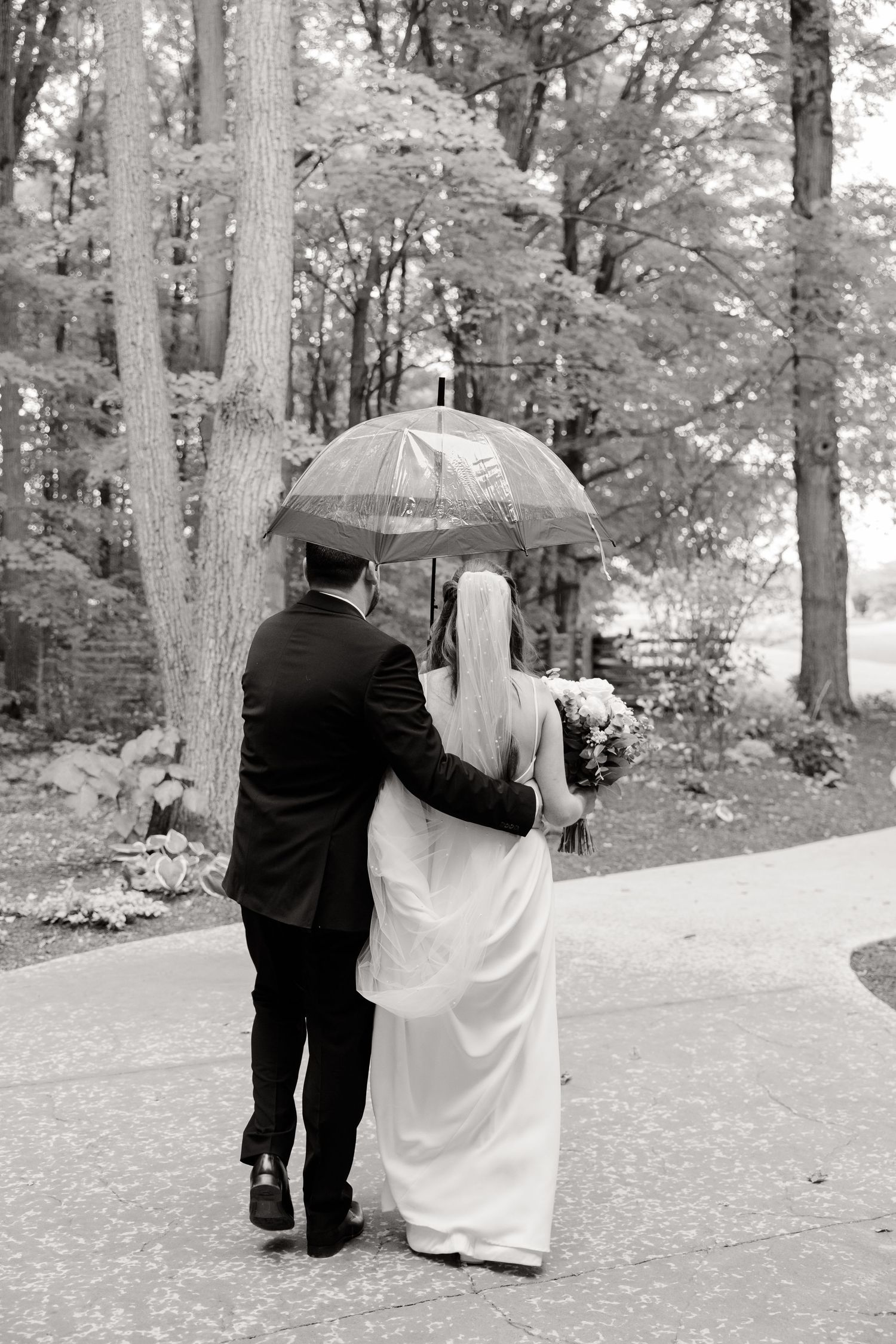 Black and white photo of couple walking with umbrella through wooded path on rainy wedding day.