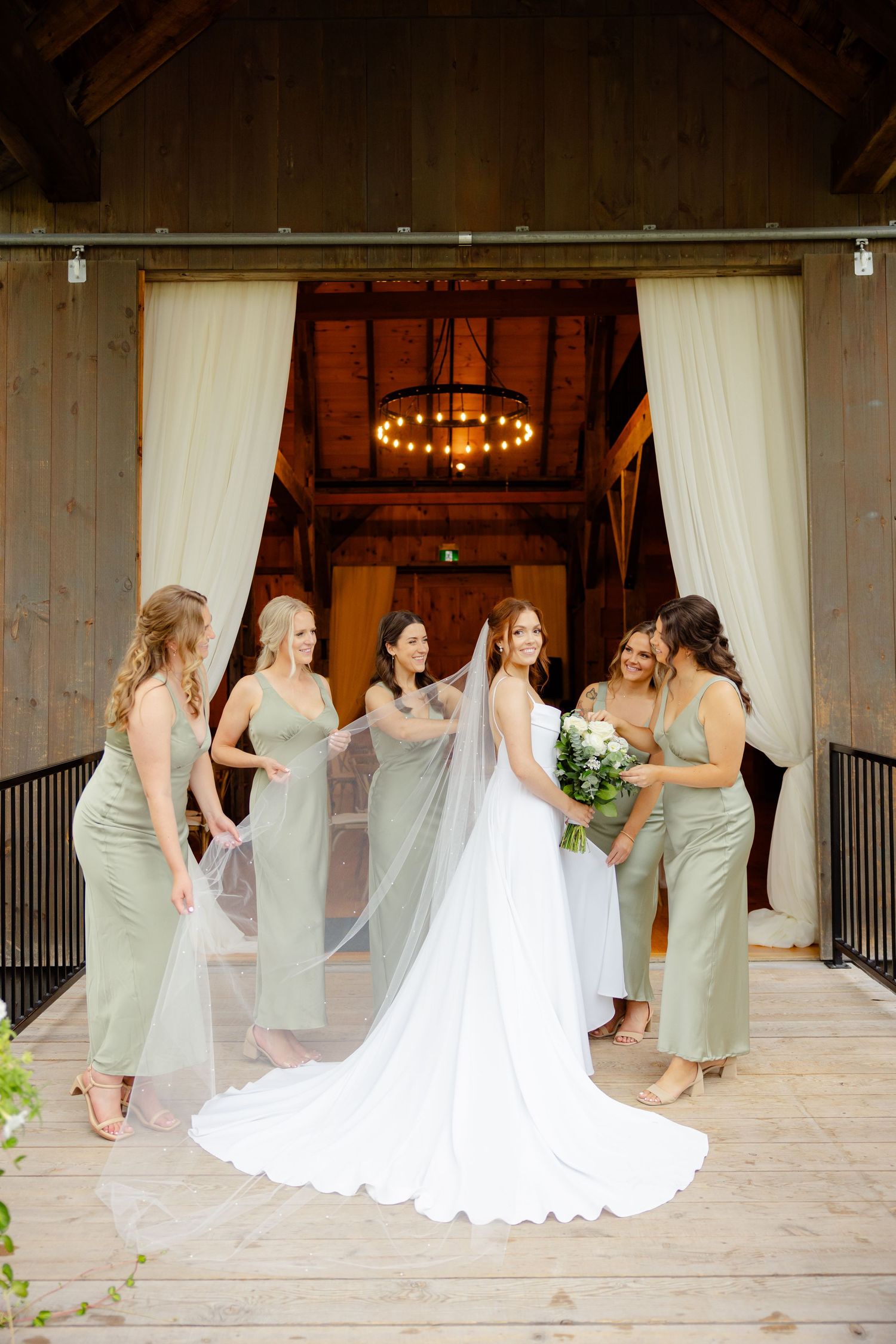 Wedding party gathering around bride in flowing white dress at rustic barn entrance with chandelier.