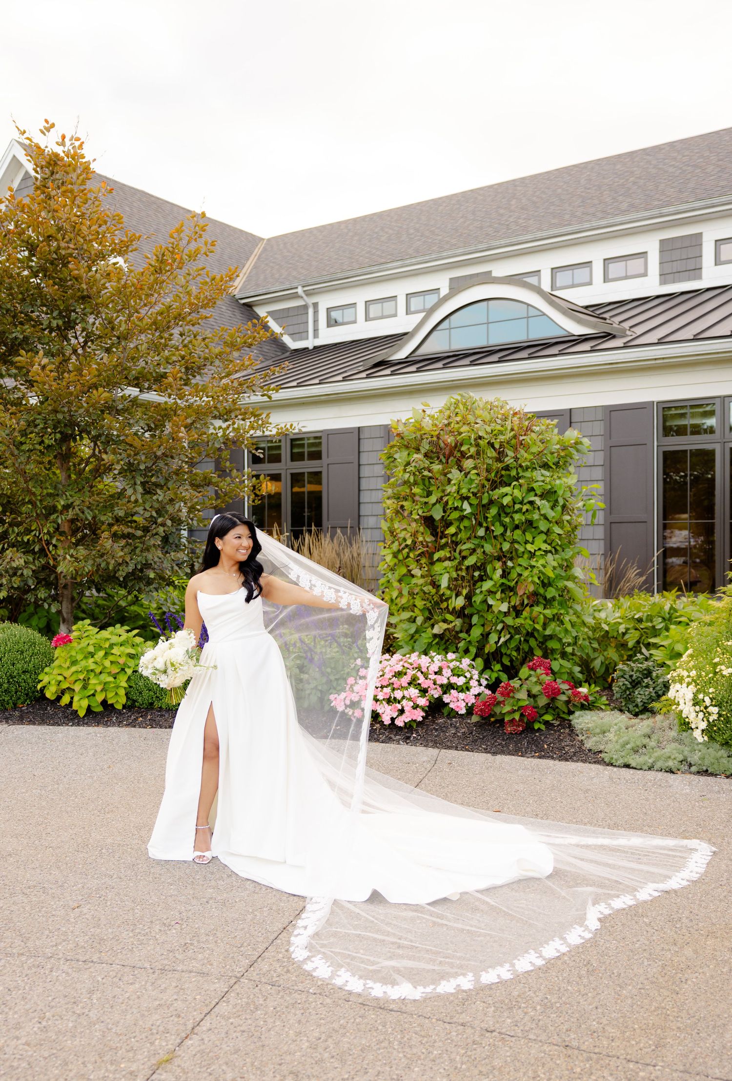 Wedding dress with dramatic cathedral veil photographed in front of grey house with autumn landscaping.