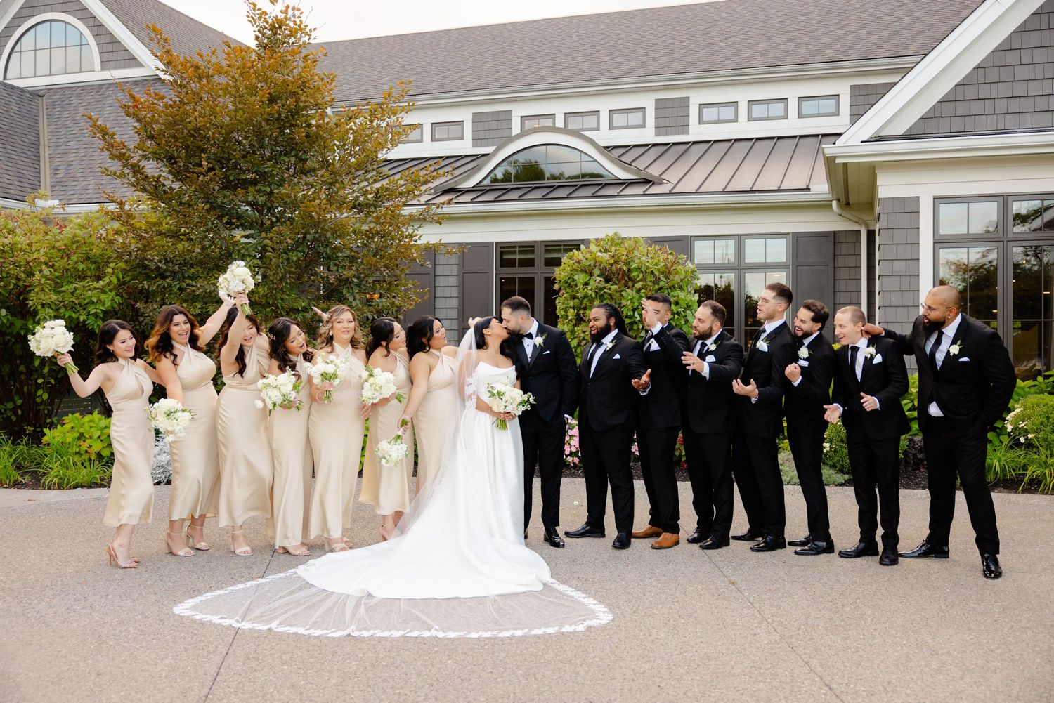 Wedding celebration group poses cheerfully outside an elegant gray building with autumn trees.
