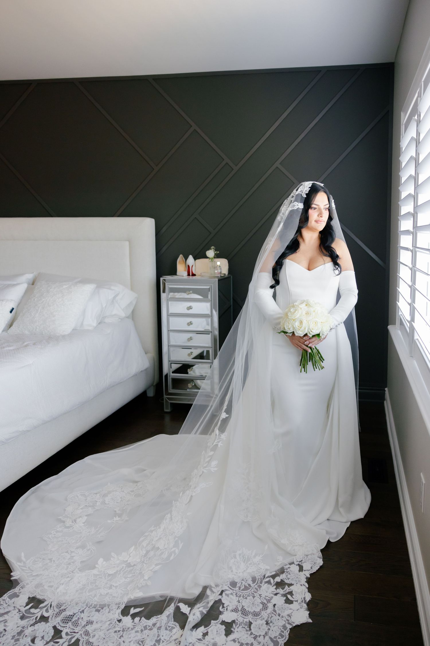 A classic bridal portrait featuring a white gown with cathedral veil against dark geometric wall decor.