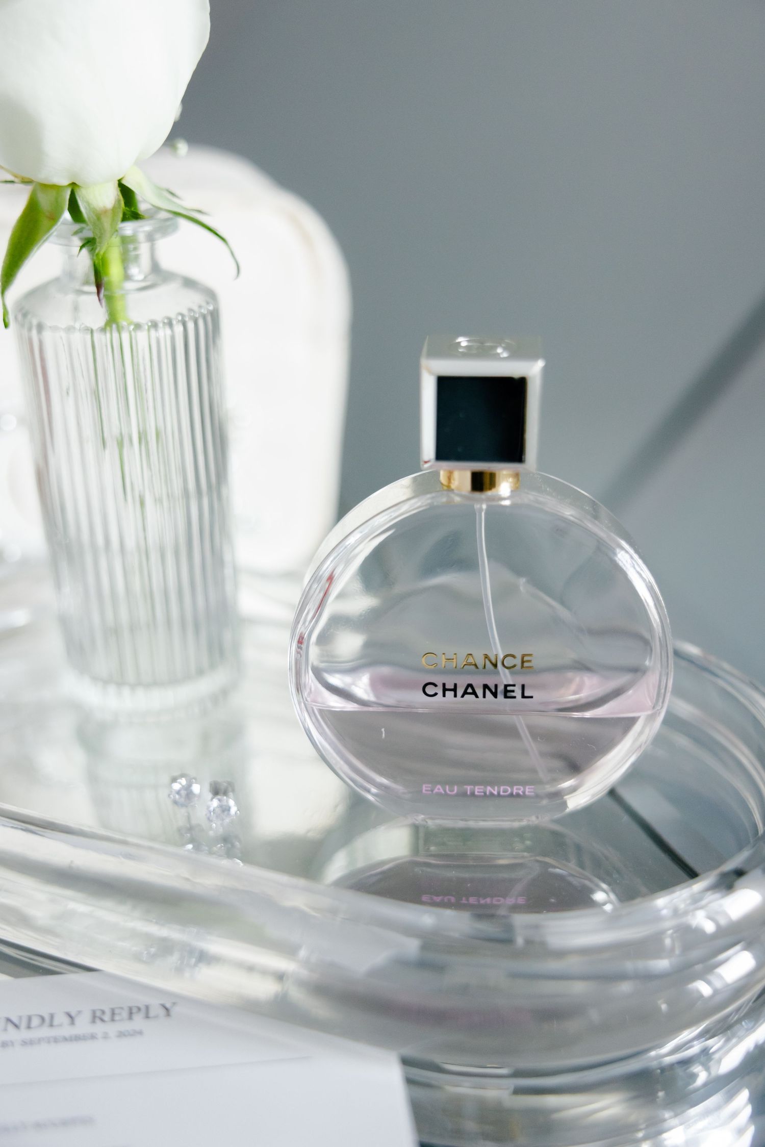 Chanel Chance perfume bottle next to white rose in glass vase.