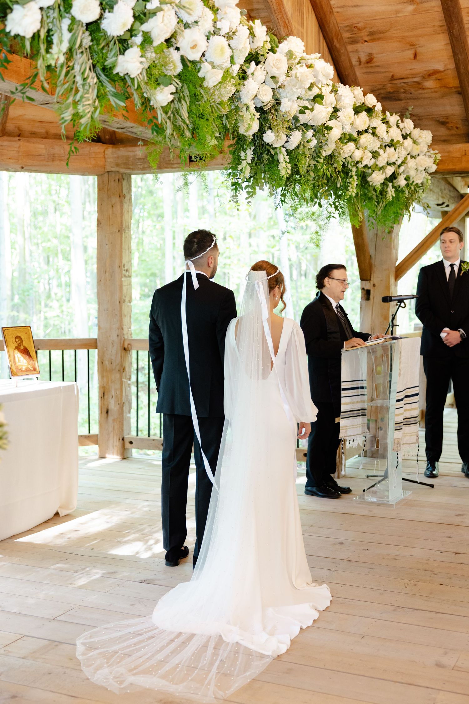 Elegant wedding ceremony in a wooden pavilion with cascading white flowers and ferns overhead.