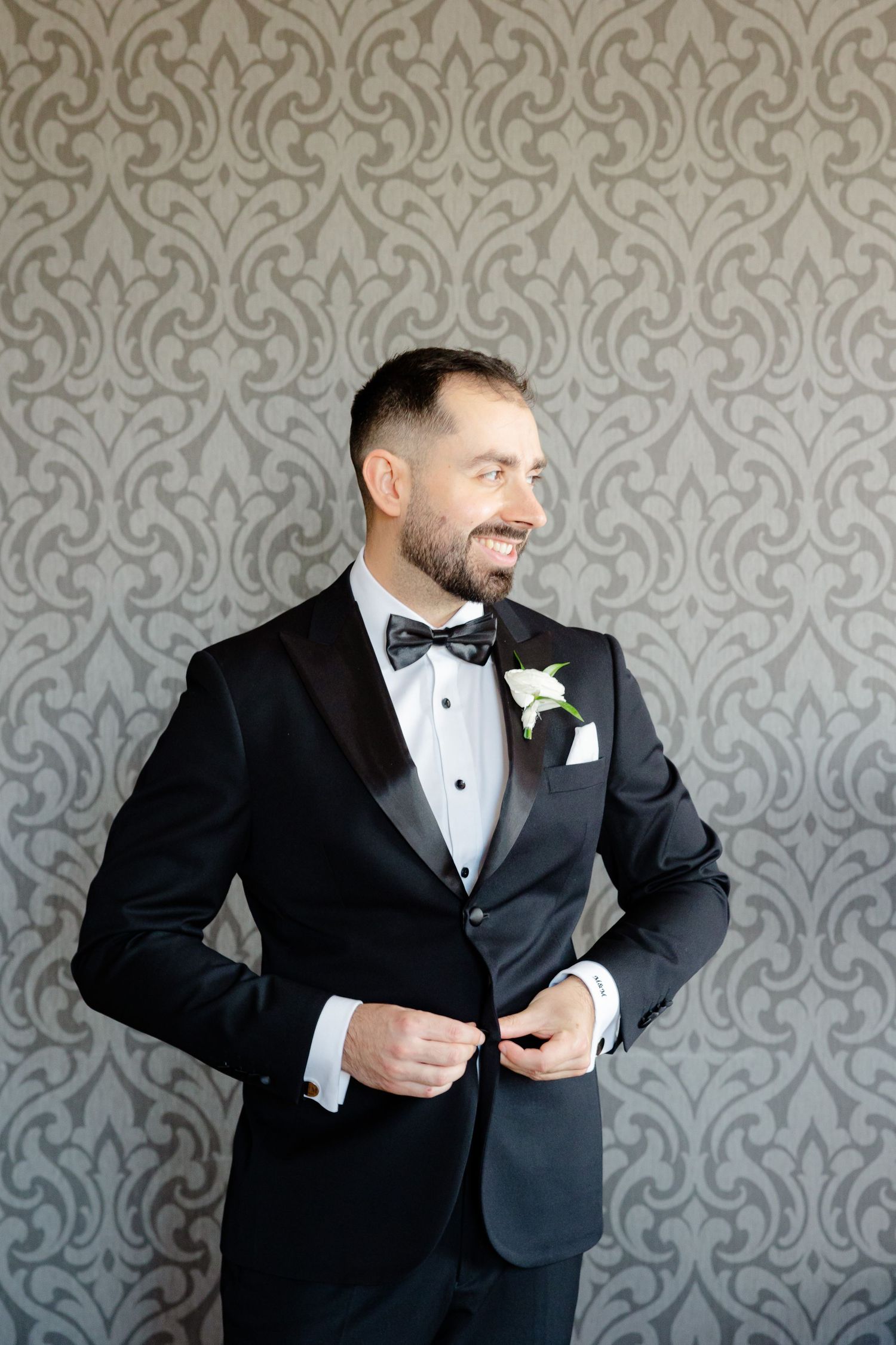 Dapper groom in black tuxedo posing against grey damask wallpaper.