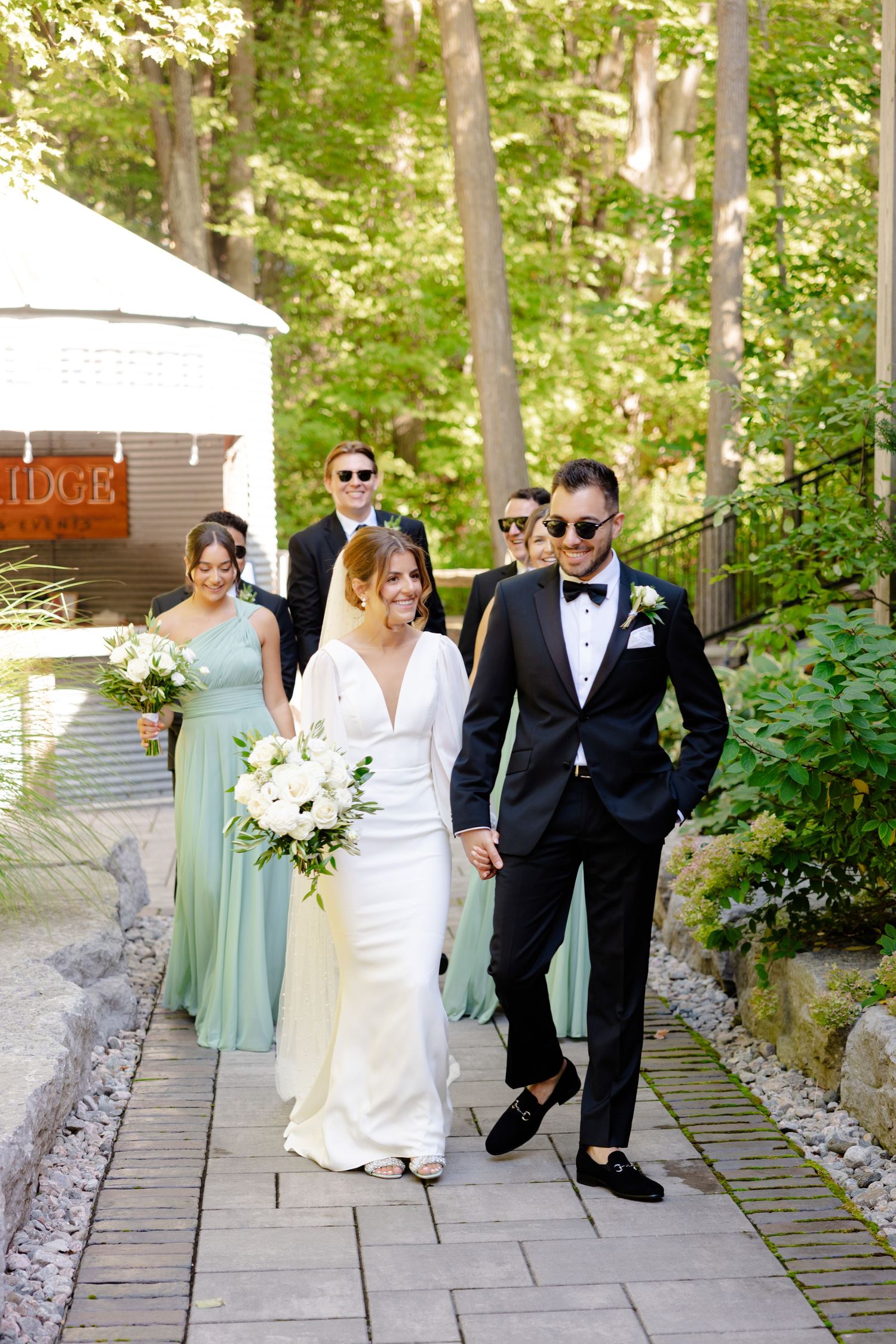 Wedding party walking along stone path with bride in white dress and bridesmaids in sage green.