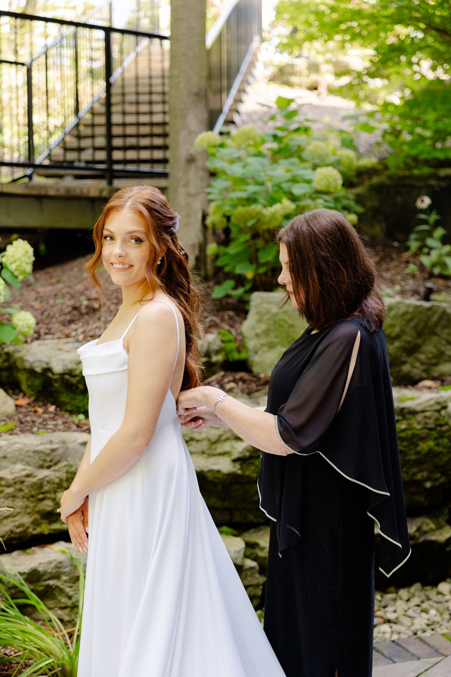 Bride in flowing white gown gets final adjustments in garden setting.