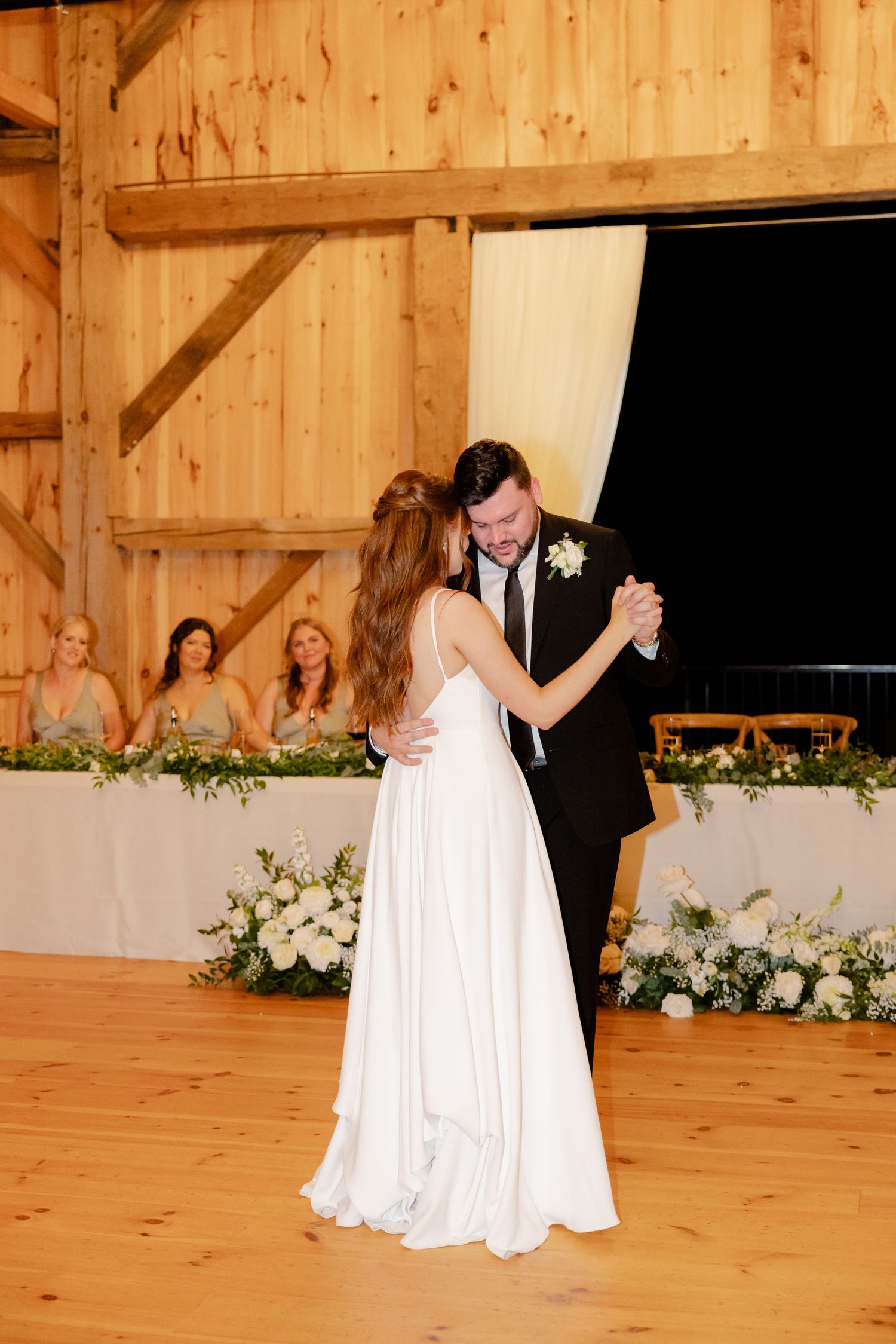 Wedding celebration first dance in rustic barn with floral decor and wedding party seated at head table.
