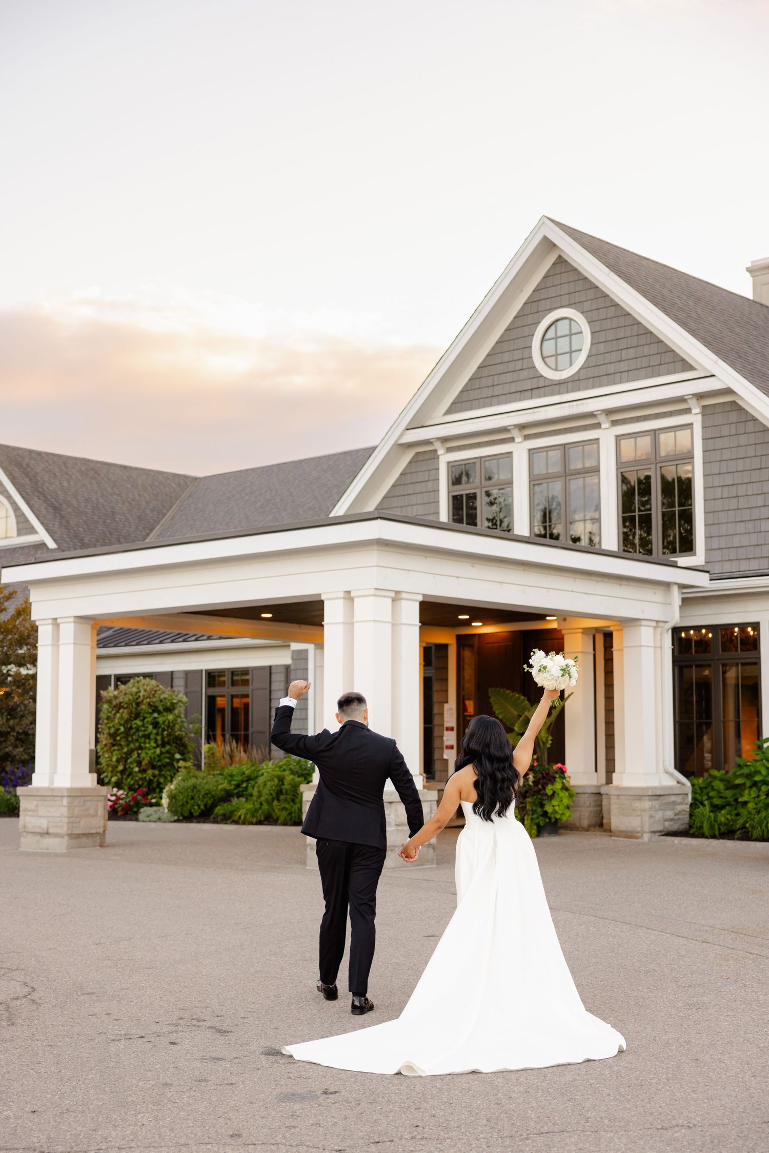 A newlywed couple walks towards a beautiful gray shingled building with white pillars.