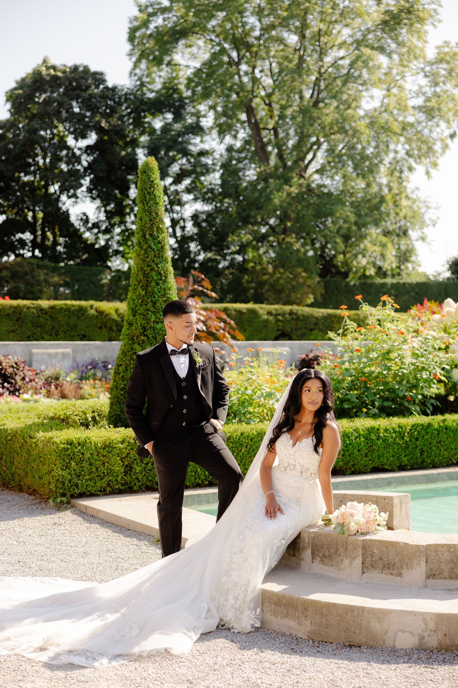 A wedding couple poses casually on stone steps in a luxurious garden setting with a reflecting pool.