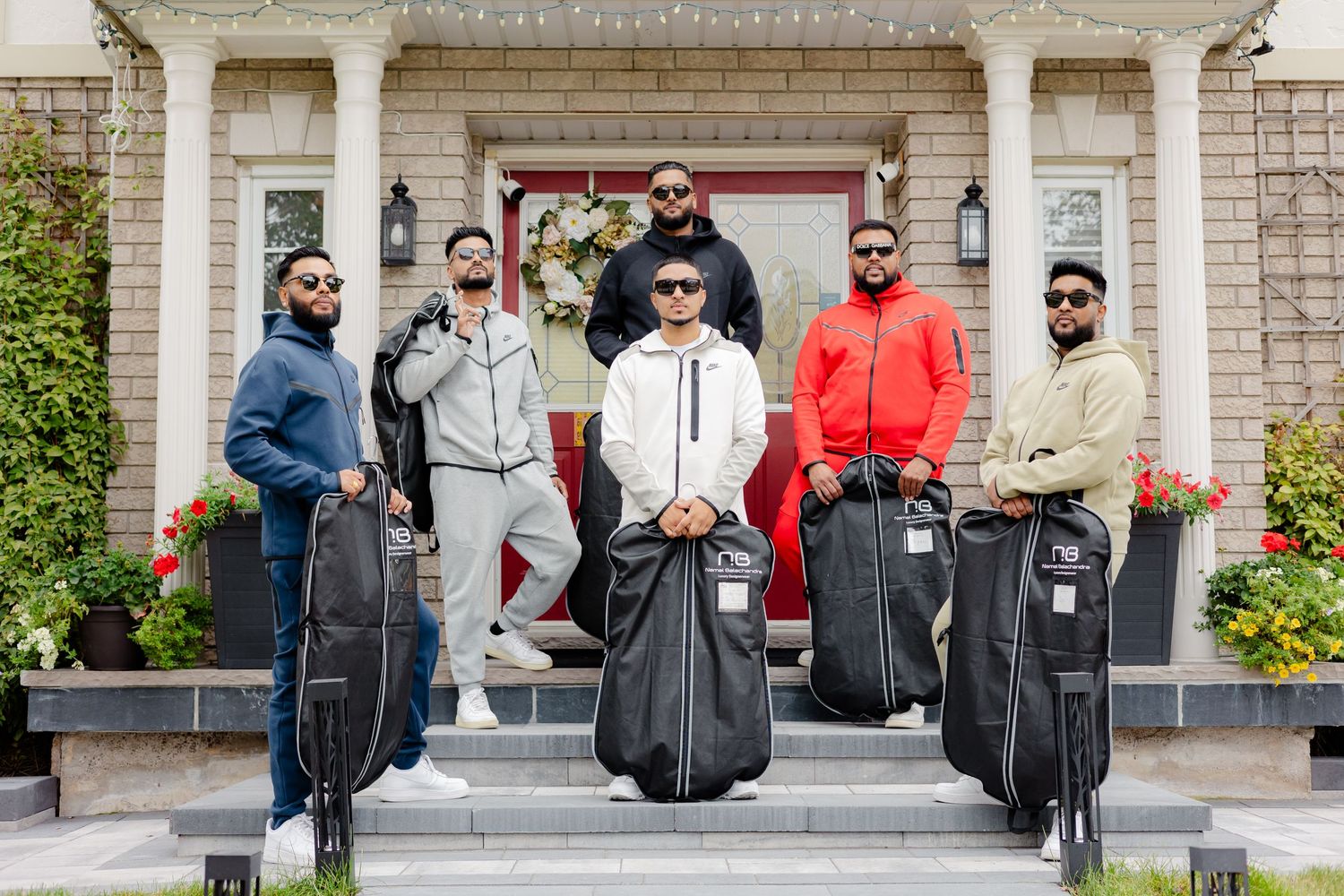 A group in casual wear poses with garment bags on brick house steps.