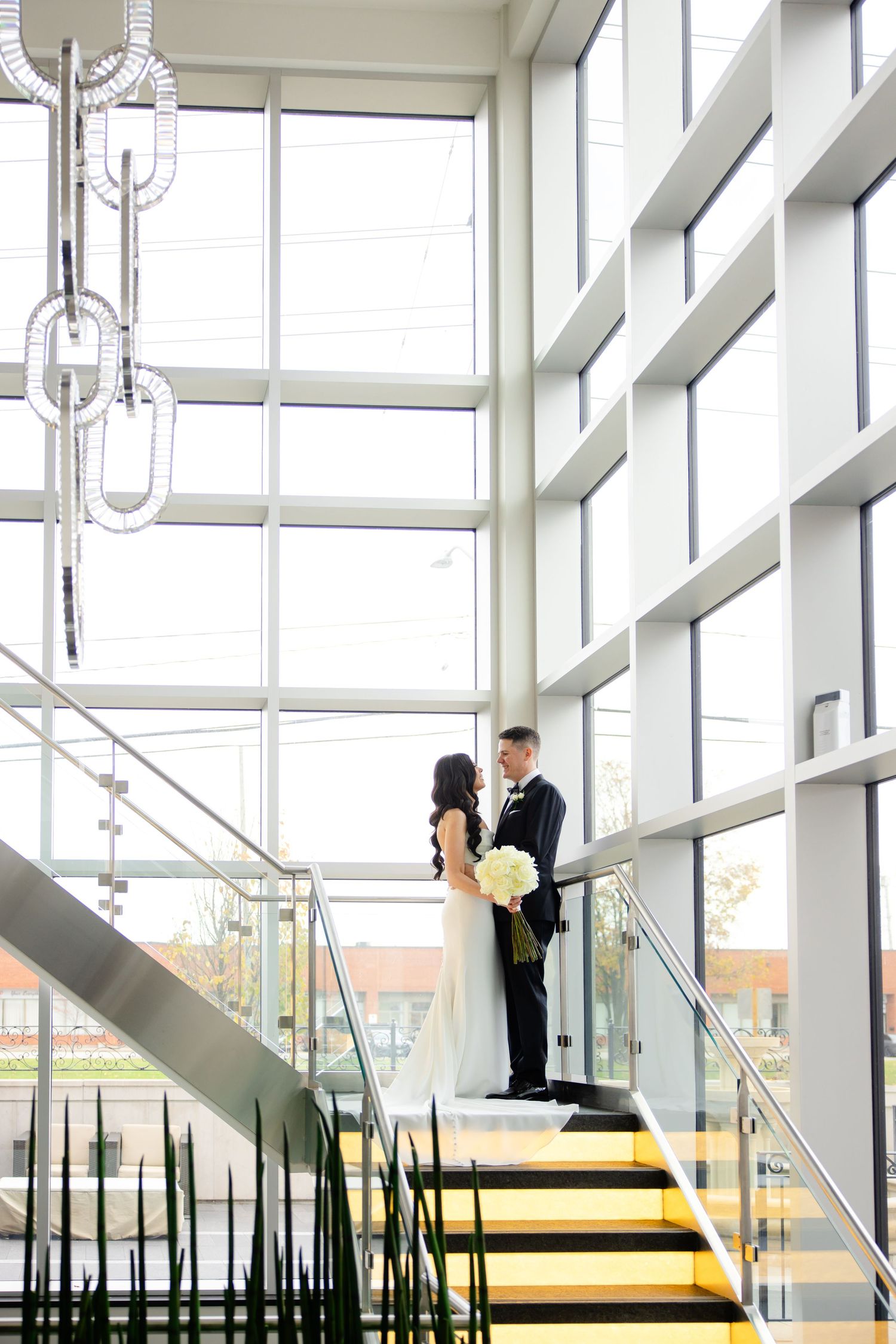 Couple in formal attire poses on bright yellow staircase surrounded by glass windows and architectural elements.