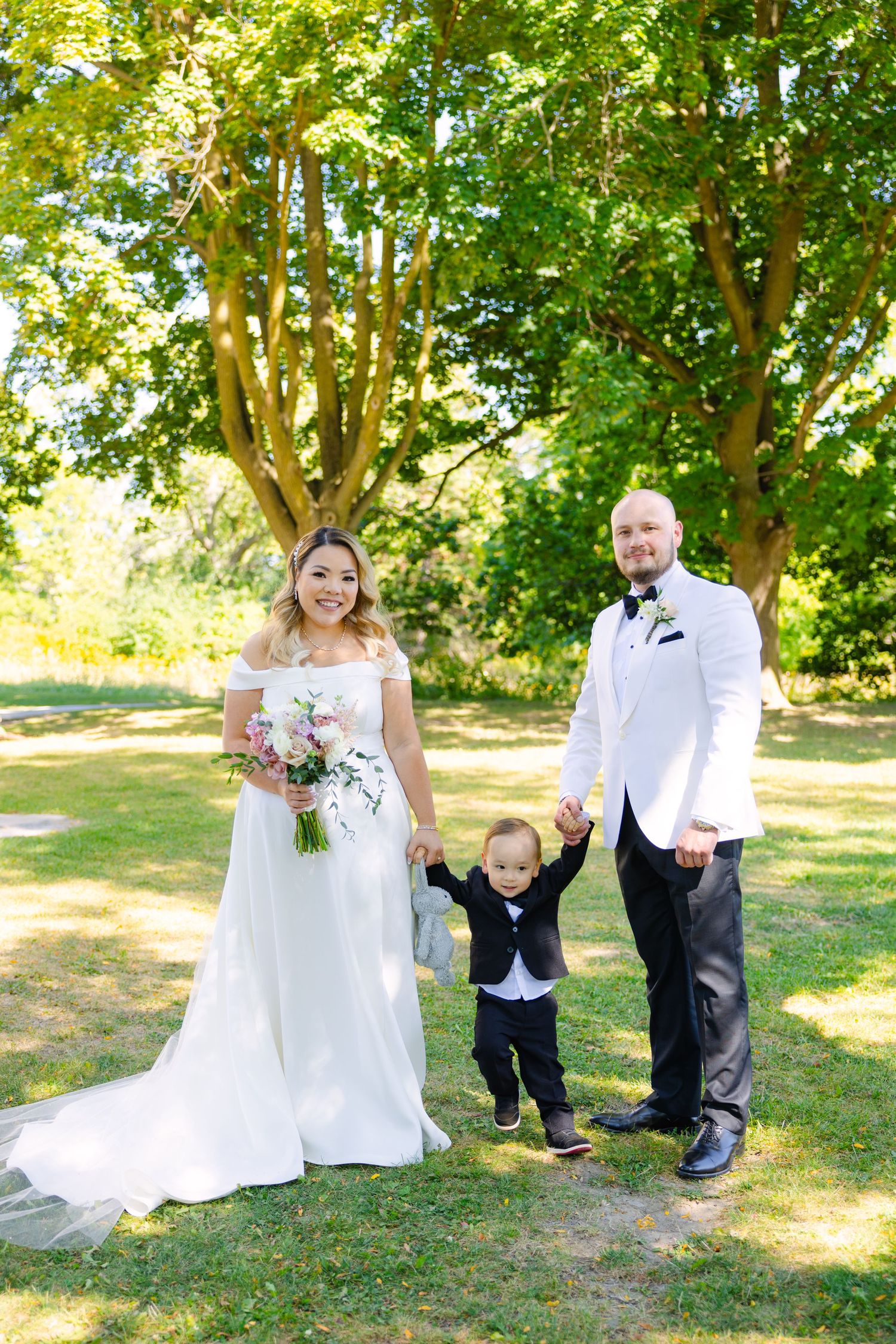 Fun family wedding photo with couple and jumping child under leafy green trees.
