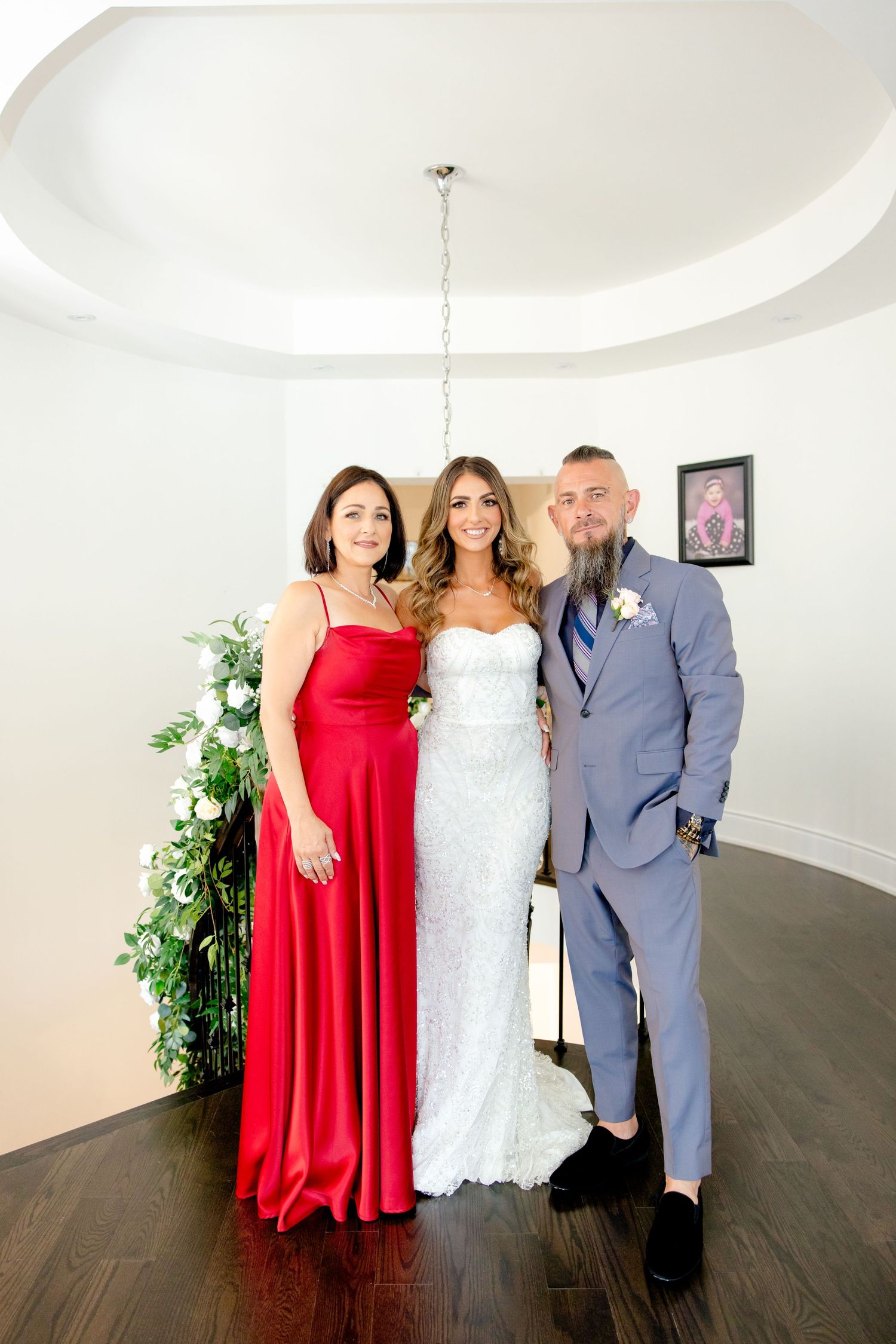 Wedding party portrait with bride in white gown alongside bridesmaid in red and groomsman in grey suit.