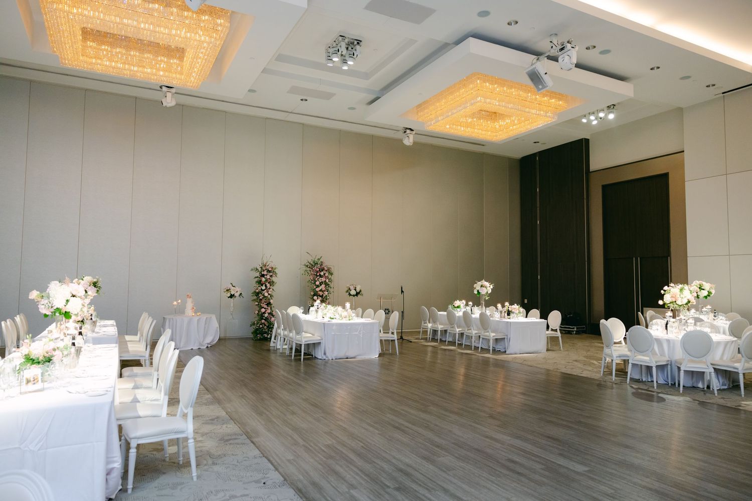 Modern wedding reception room with white chairs and gold chandeliers on a wood floor.