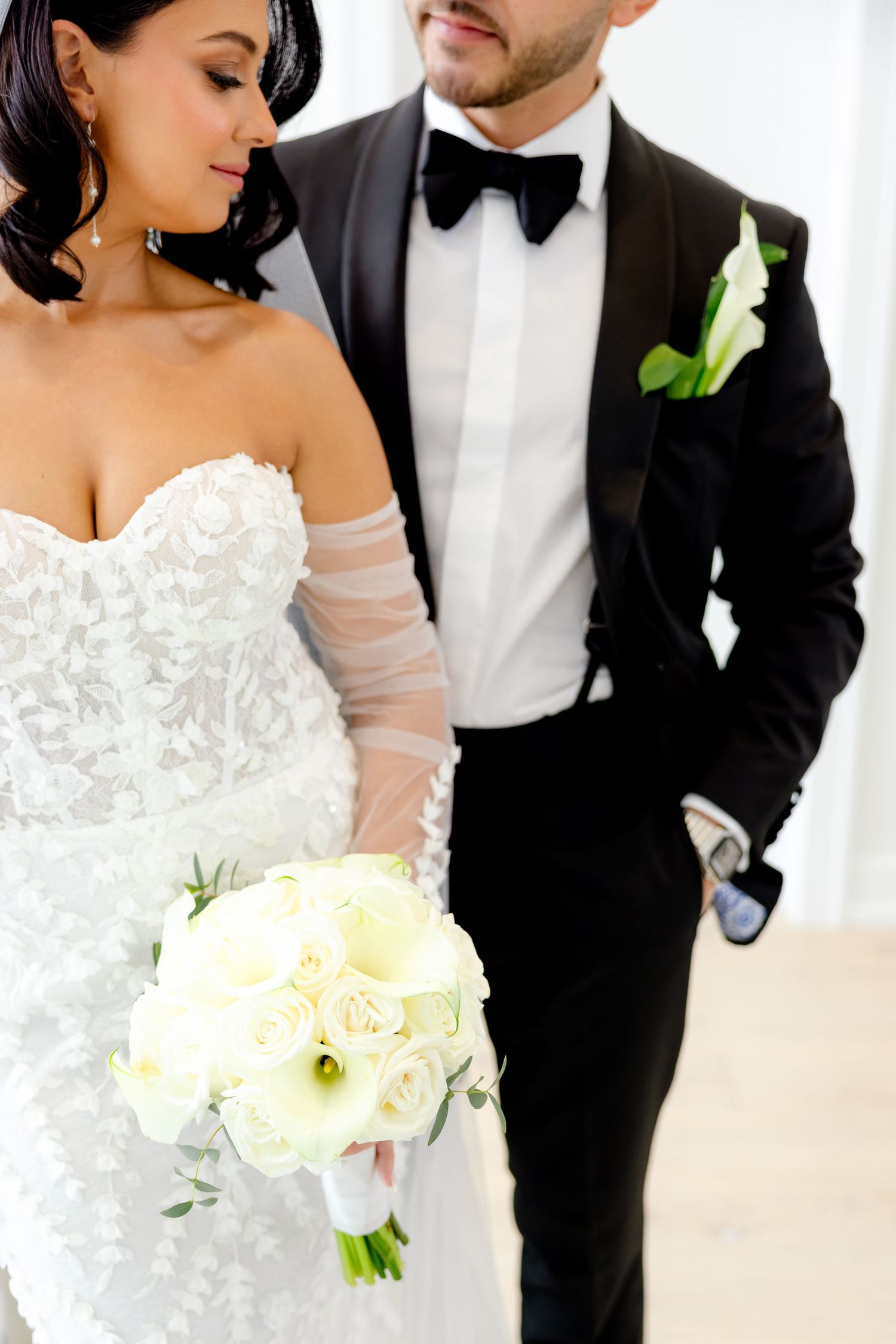 A bride in a lace gown holds a white bouquet while standing next to her groom in a classic black tuxedo.