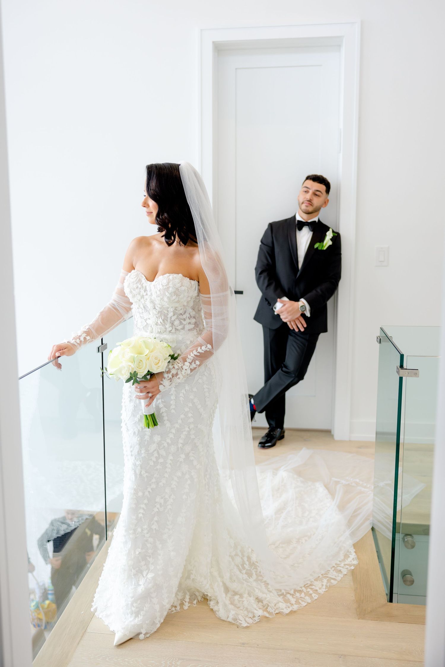 Bride holds bouquet while groom leans against doorframe in modern hallway.