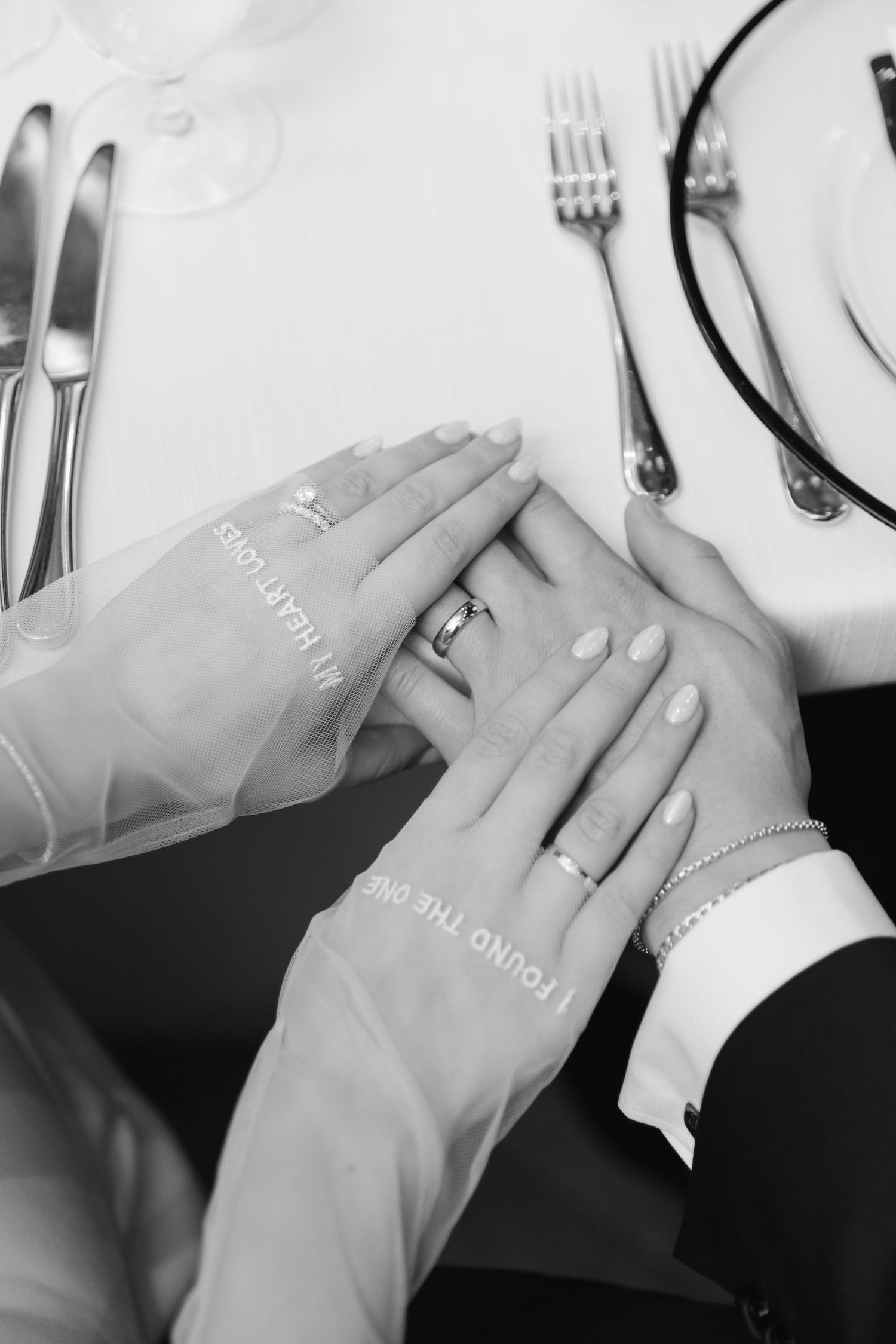 Black and white photo of newlywed hands with rings on an elegant dining table.