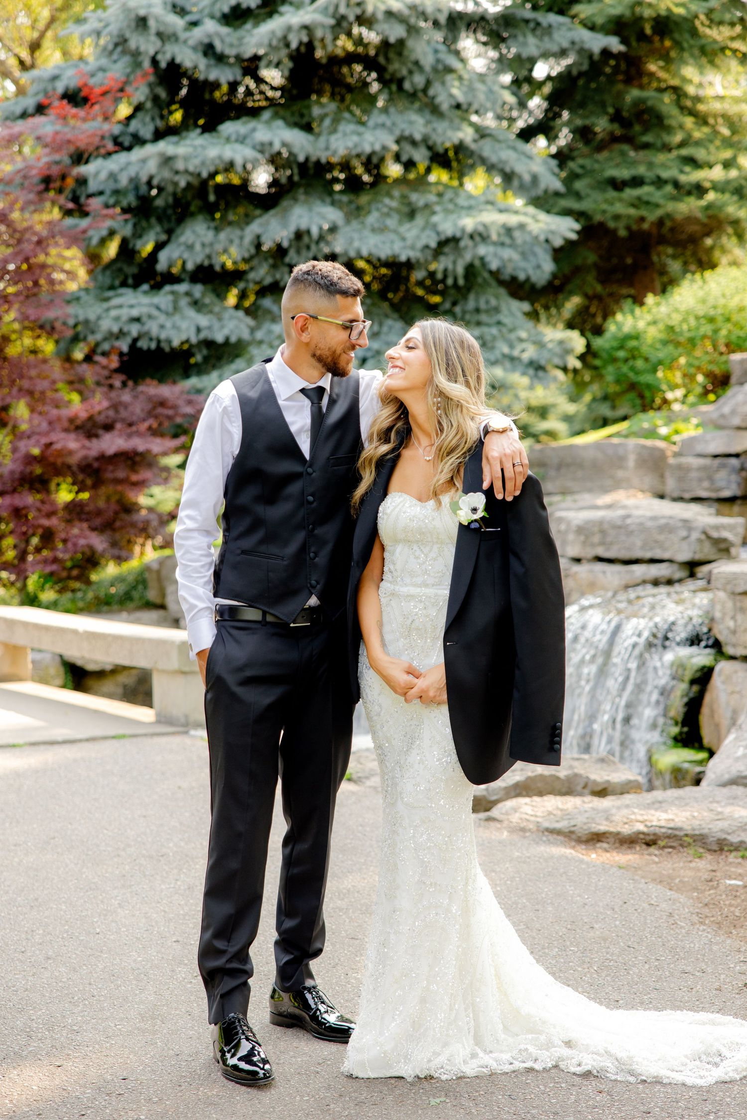 Newlyweds share an intimate moment by a water feature with blue spruce trees in background.