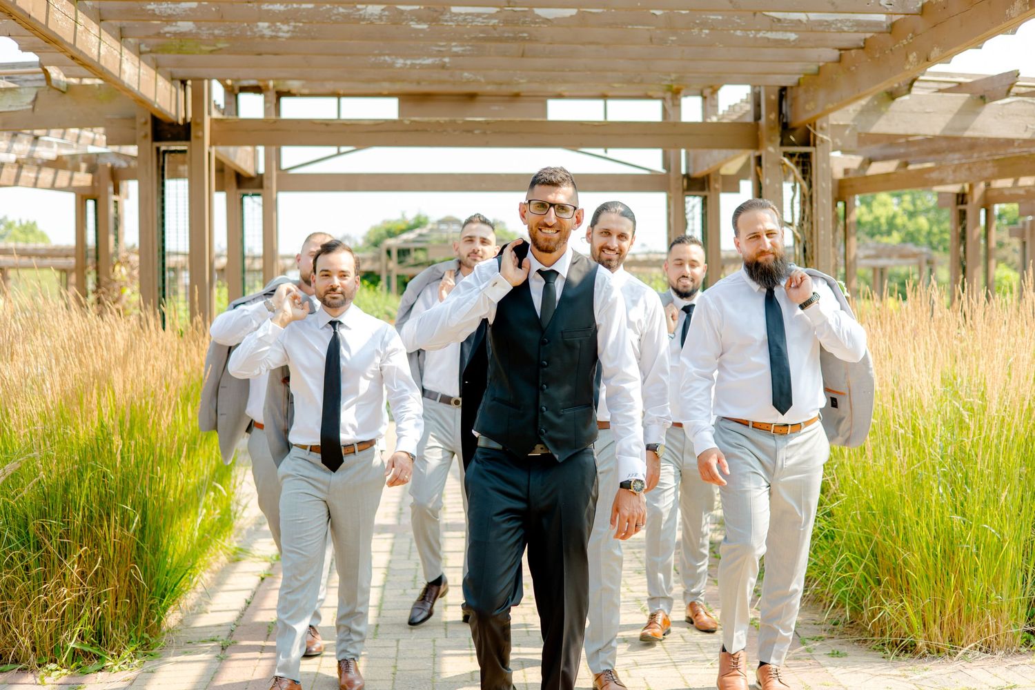 Groomsmen in light suits walking with groom in navy suit through outdoor garden pathway.