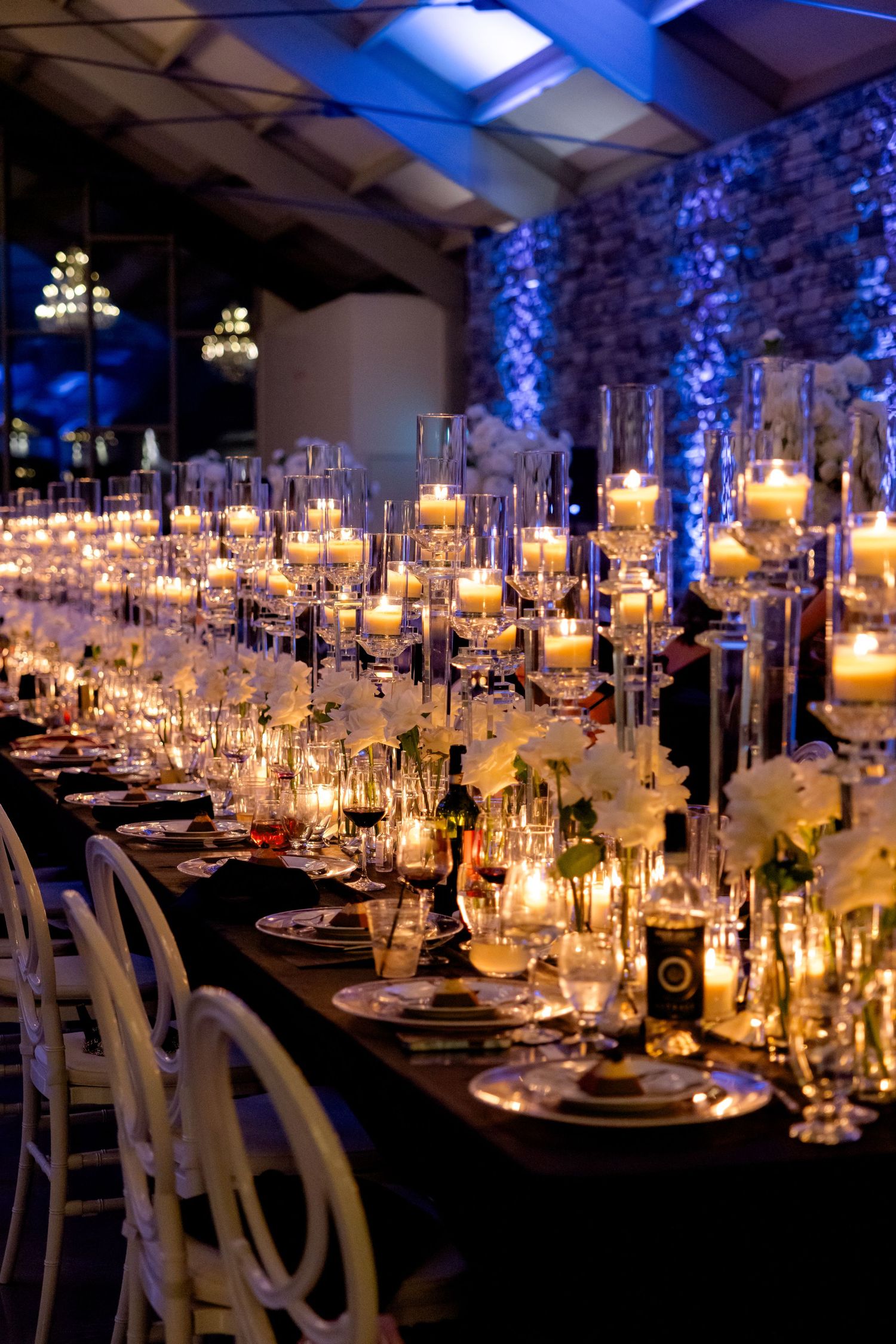 Elegant wedding reception table with glowing candles and place settings under romantic blue uplighting.
