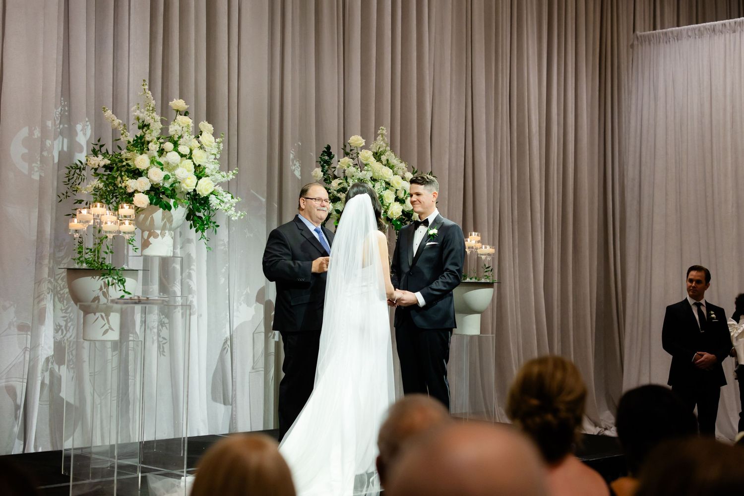 Wedding ceremony taking place on stage with white floral arrangements and sheer fabric backdrop.