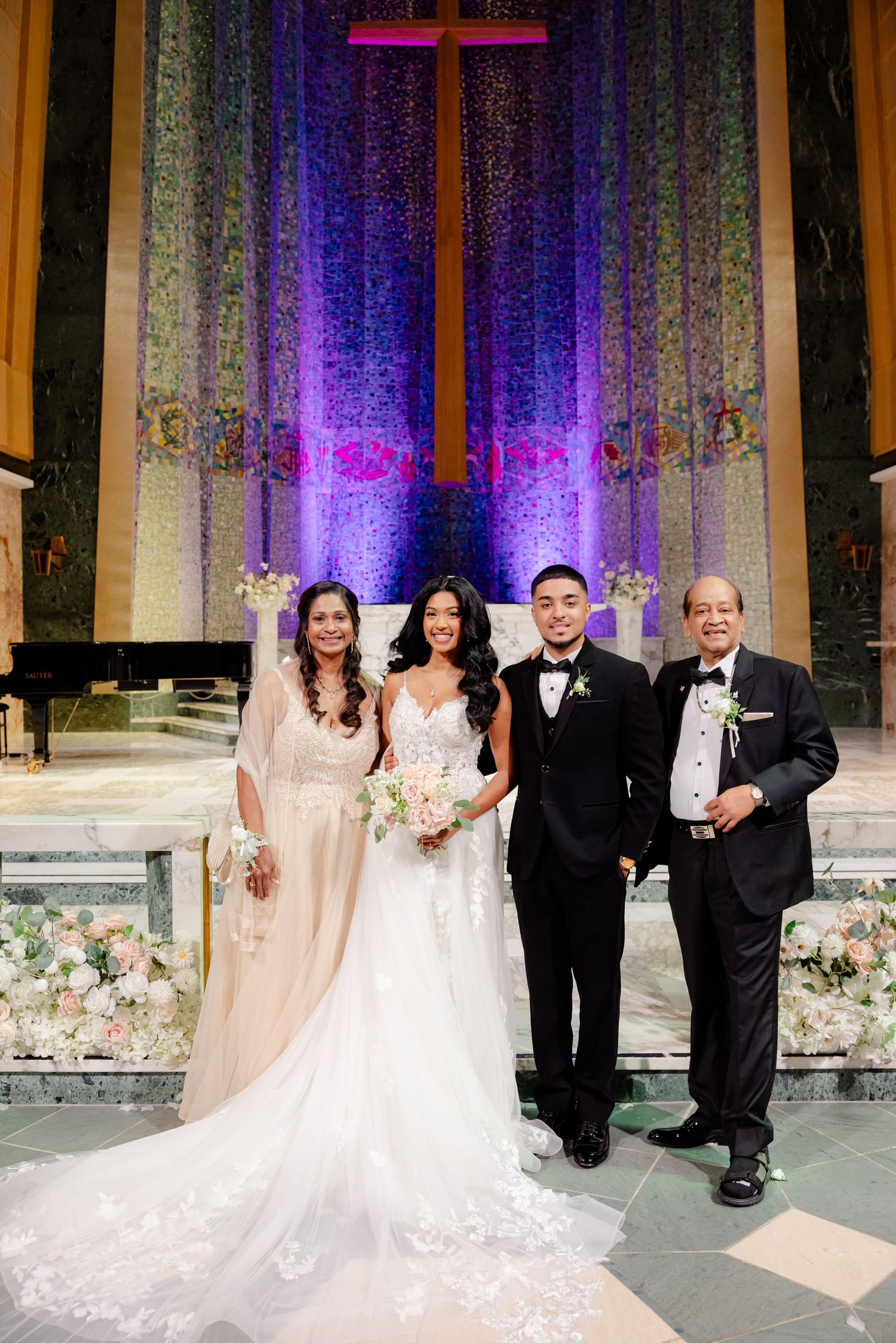 Wedding party poses in front of purple-lit church altar with cross and floral decorations.