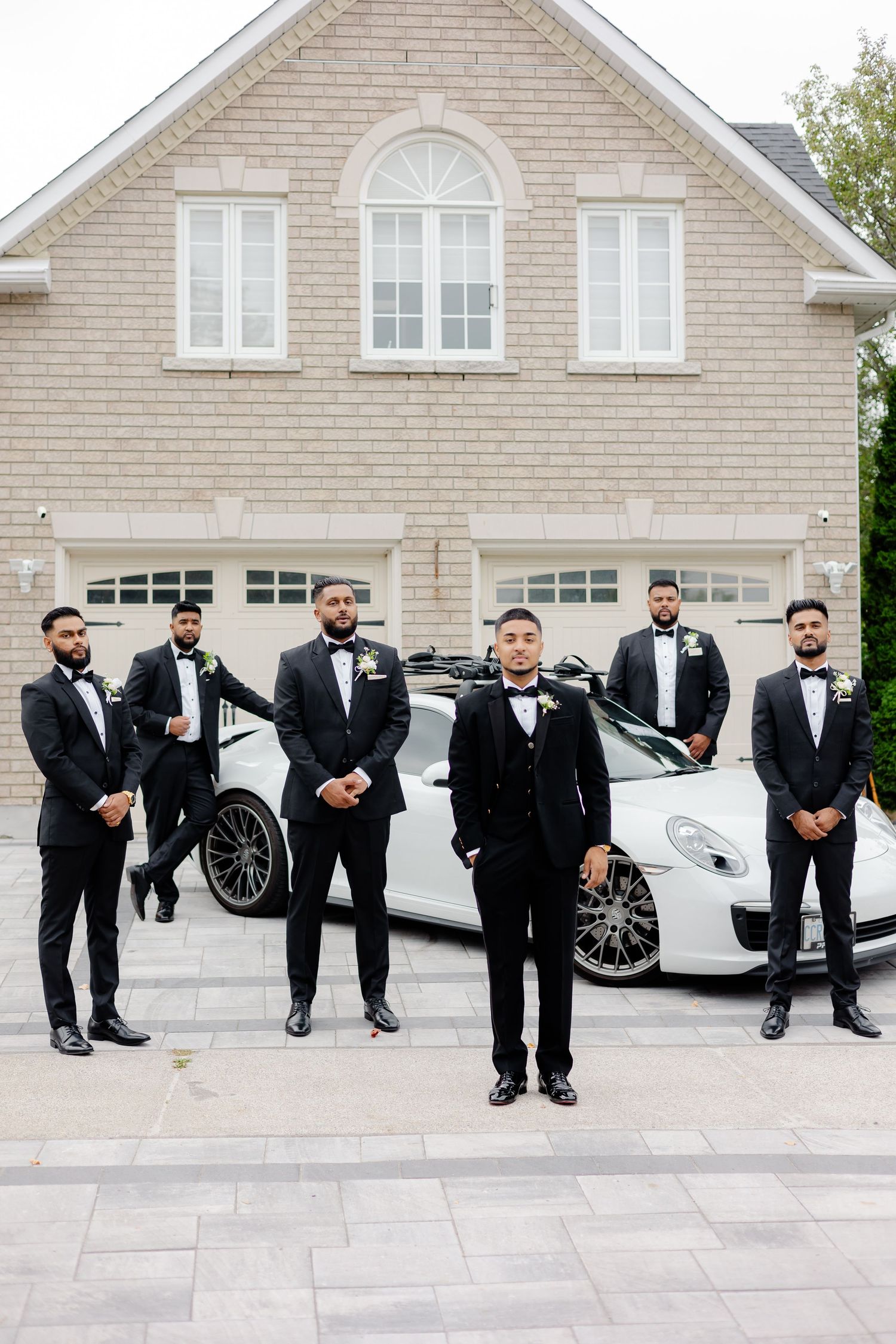 Wedding party in formal attire stands confidently beside luxury sports car in front of garage.