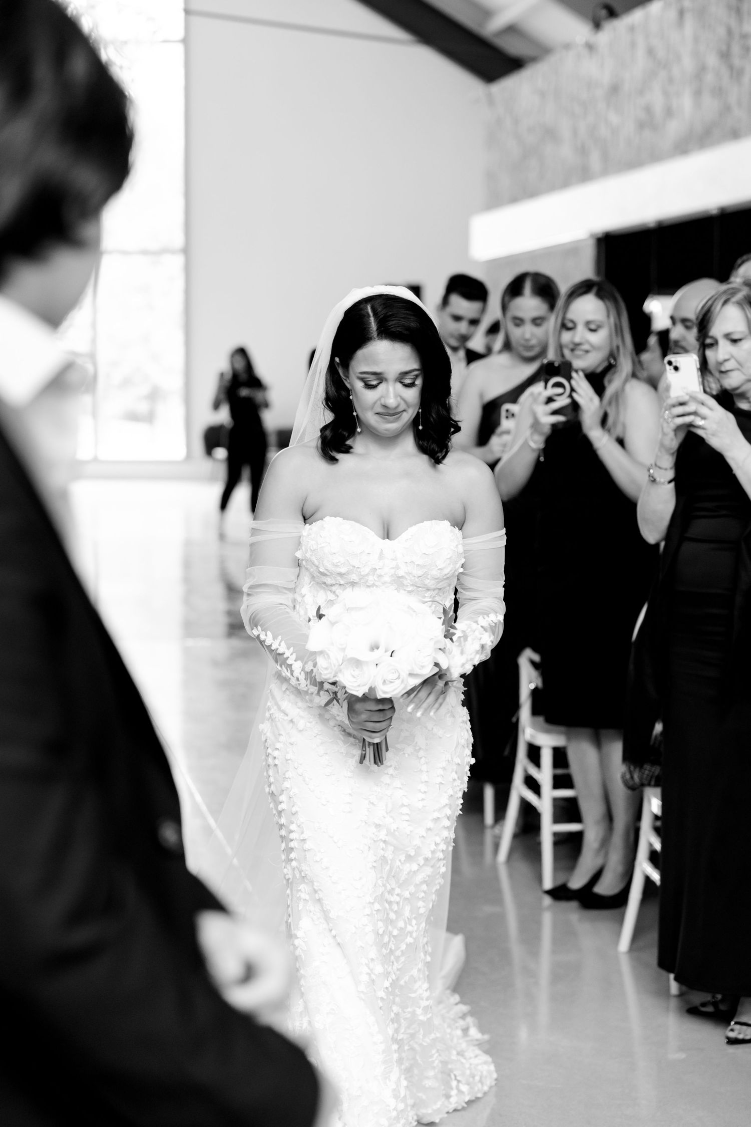 A bride in a fitted lace wedding dress walks down the aisle holding a white bouquet as guests look on.