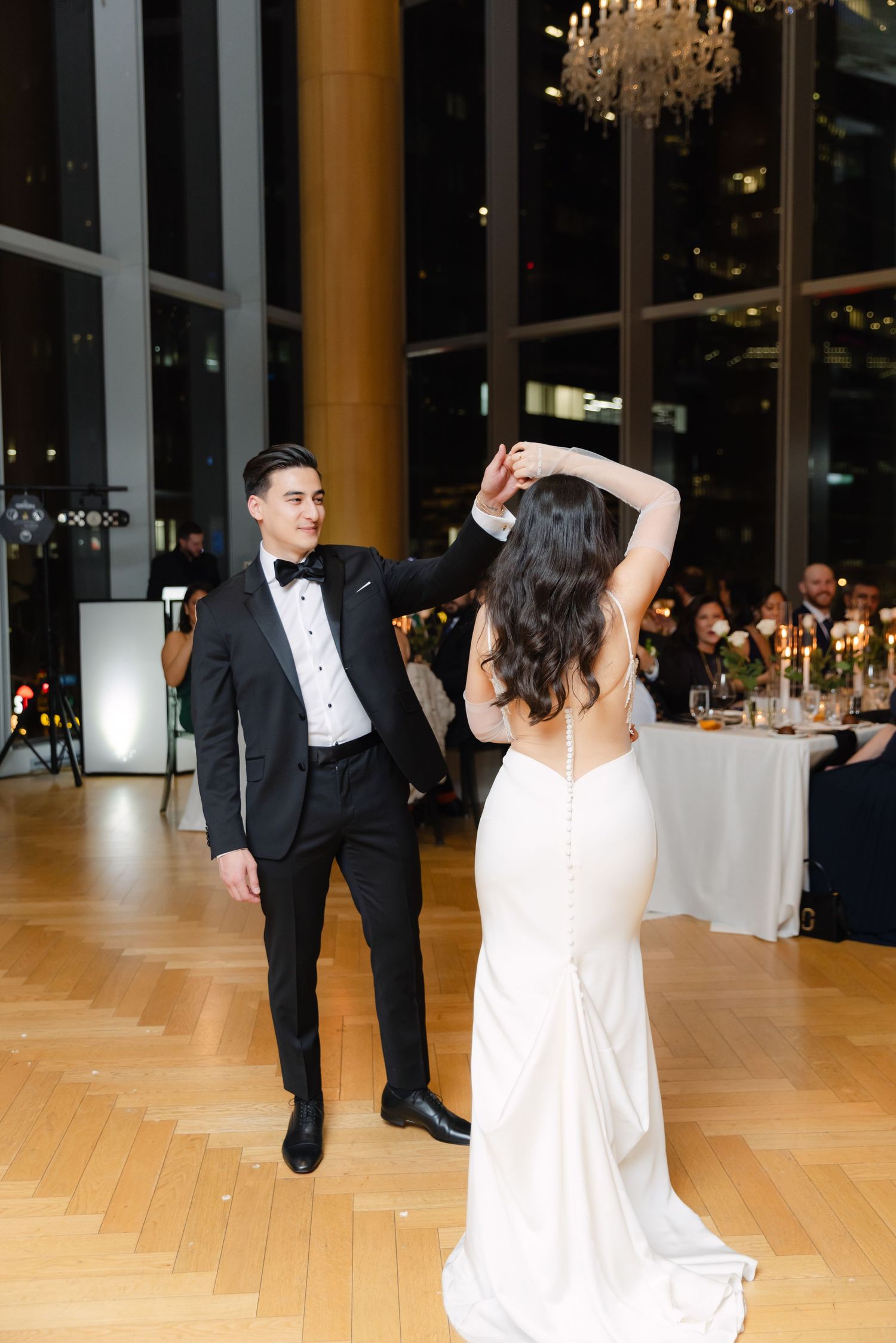 Elegant couple performs first dance on parquet floor with chandeliers and reception tables in background.