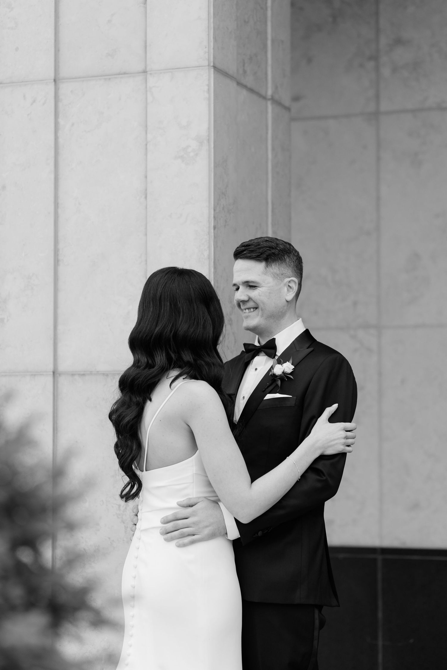Black and white intimate wedding portrait of couple embracing against stone column backdrop.