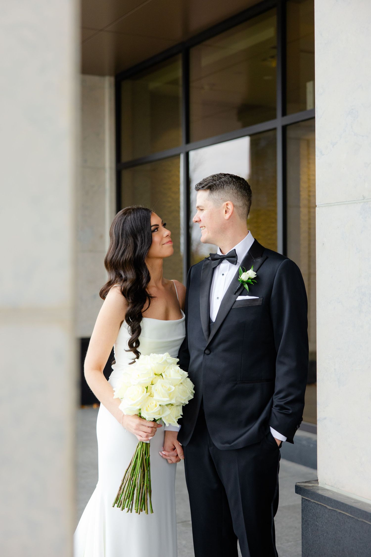 A couple in wedding attire poses with a white bouquet against modern architecture.
