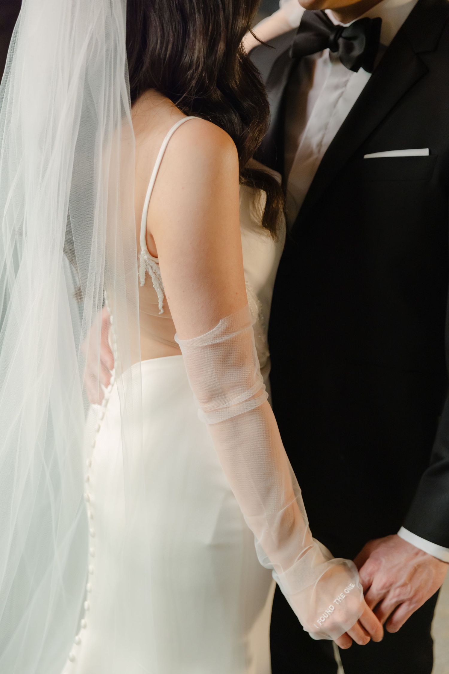 Close-up detail of wedding attire showing long white gloves and classic black tuxedo.
