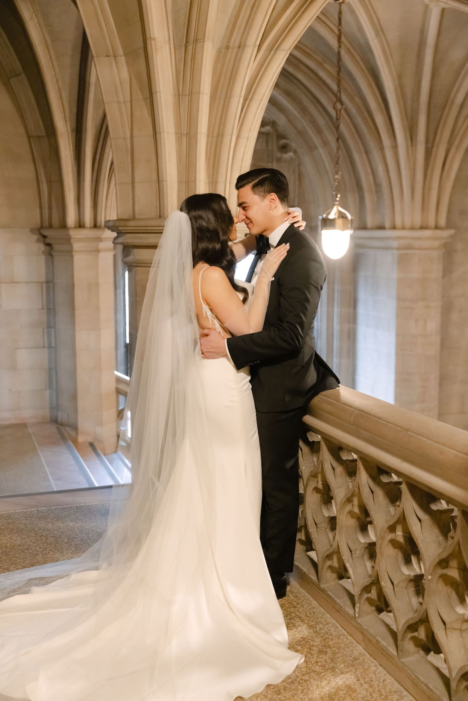 Bride in flowing white gown and groom share intimate moment in Gothic hallway.