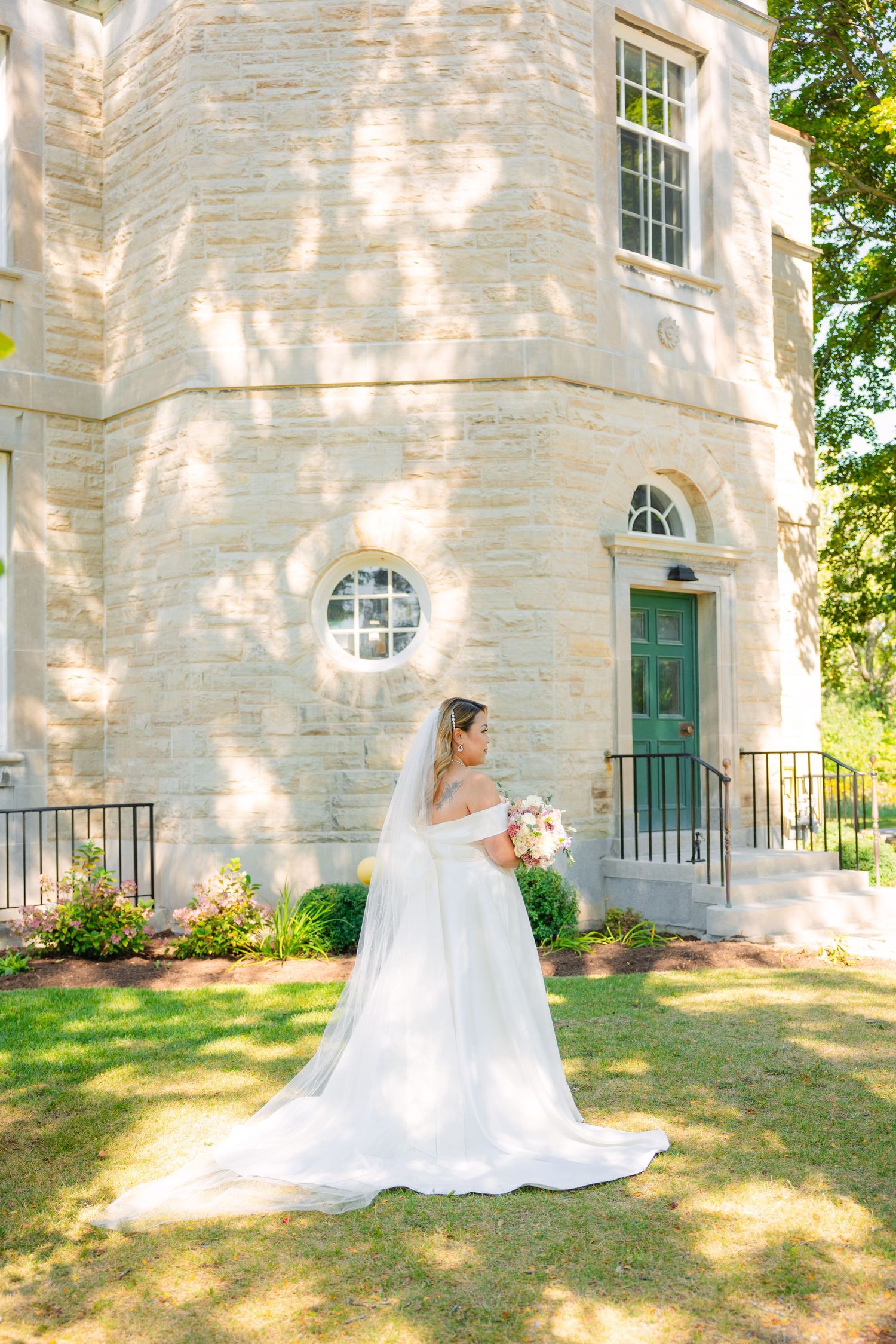 Bride in classic white dress poses in front of stone building with round window and green door.