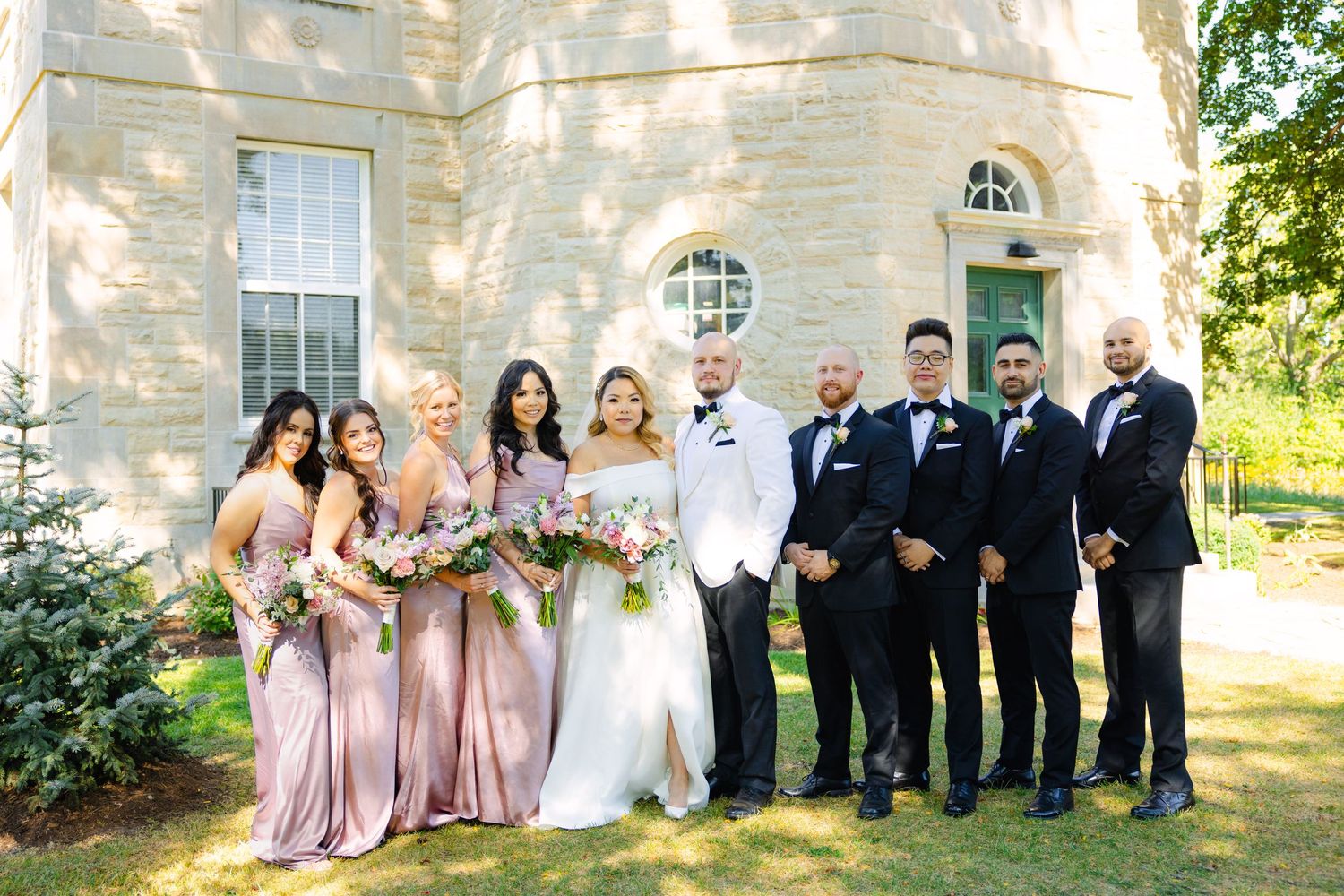 Wedding party in pink dresses and black tuxedos pose together against stone building with round windows.