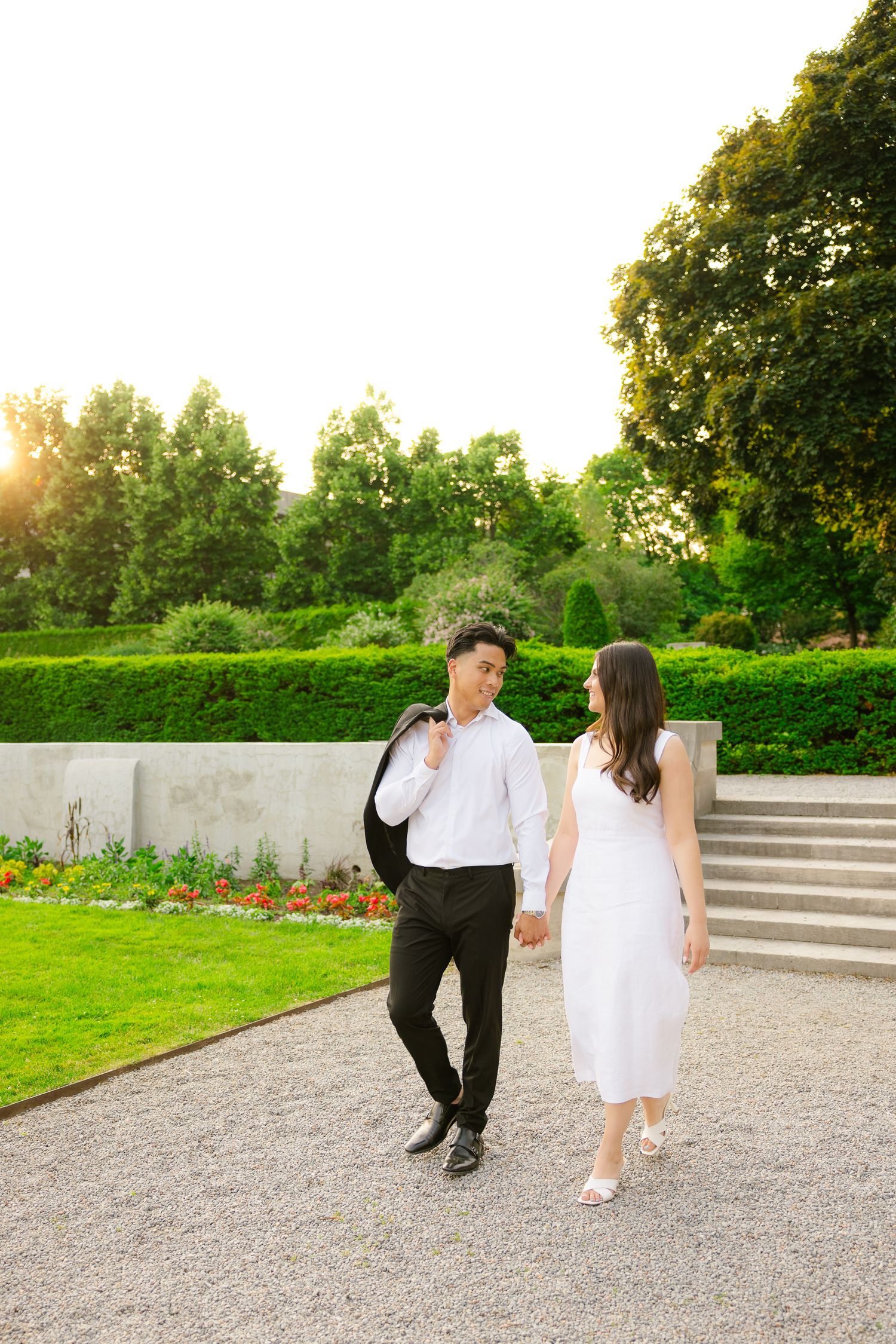 A couple walks hand in hand on a gravel path with flowering gardens and stone steps behind them.